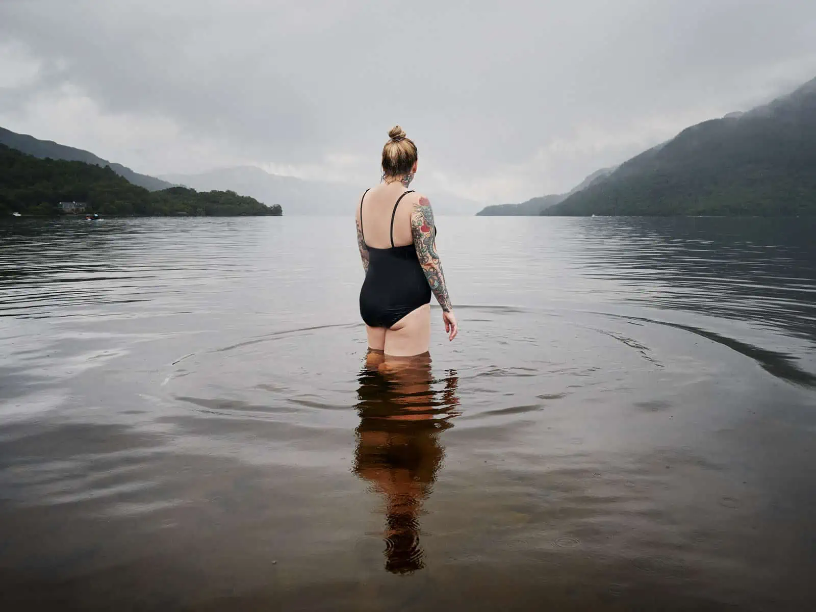 ID: A landscape image. Fay stands with back to the camera in a black swimsuit in a loch. It is misty and moody and quite dark. There are ripples forming circles around Fay's legs. ⁣