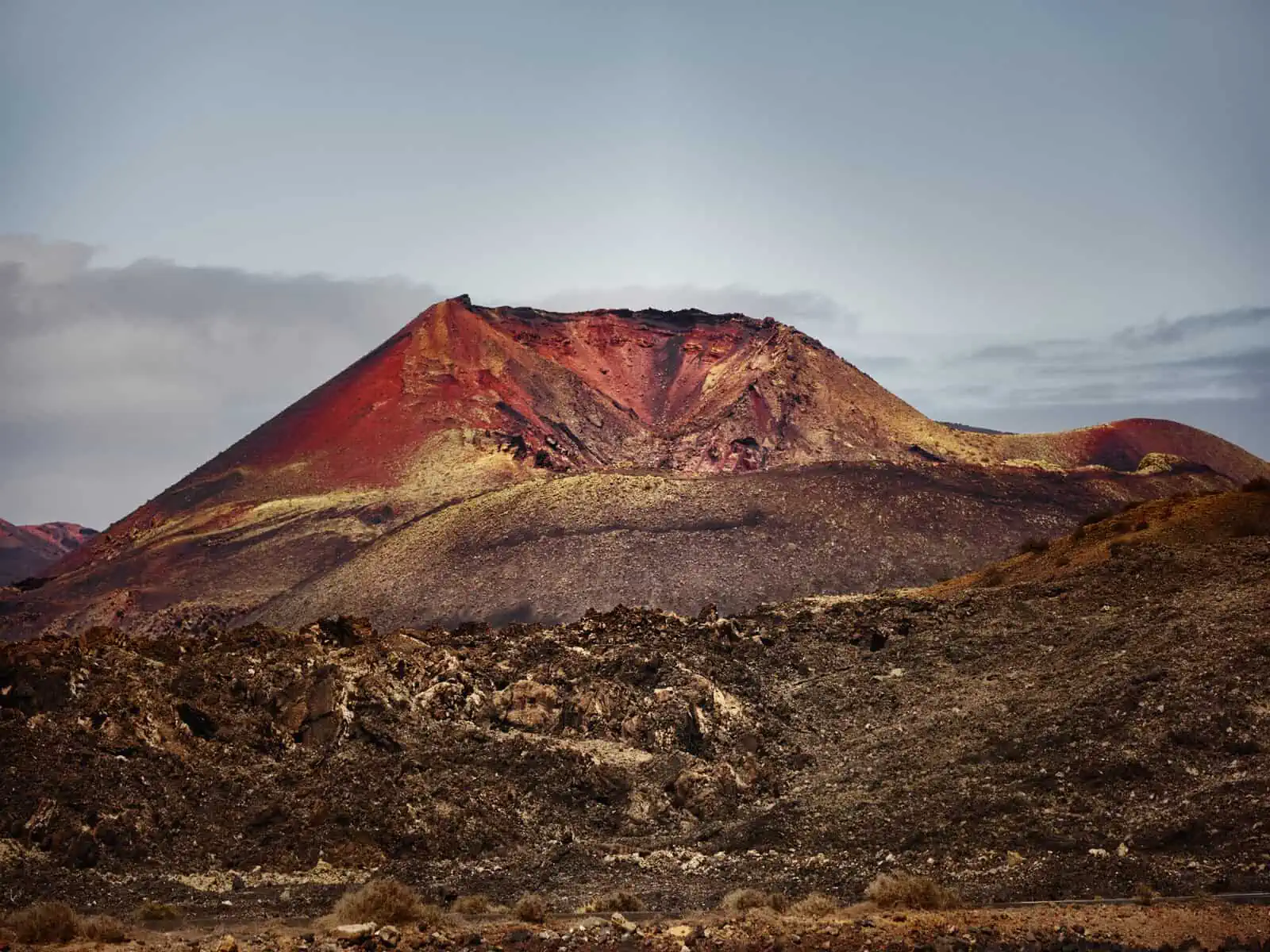 Image description: A landscape image. The foreground is framed with volcanic rubble - in deep browns and covered in green lichen. The background of the image is a volcano which is many colours of red, orange and yellow. The sky is blue with backlit …