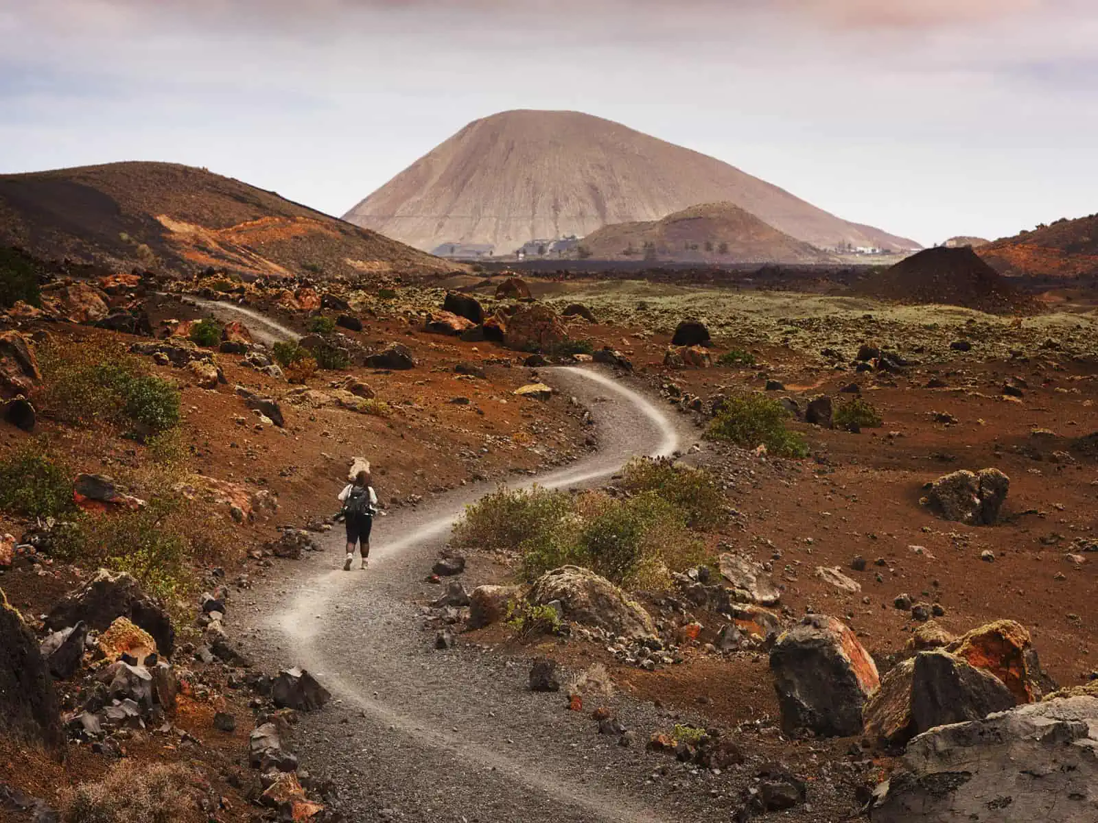 𝘐𝘮𝘢𝘨𝘦 𝘥𝘦𝘴𝘤𝘳𝘪𝘱𝘵𝘪𝘰𝘯: Landscape image. Fay is walking along a path through a volcanic, mountainous landscape with her back to the camera. There are many earthy colours that make up the scene - such as browns, reds and oranges. It is sli…