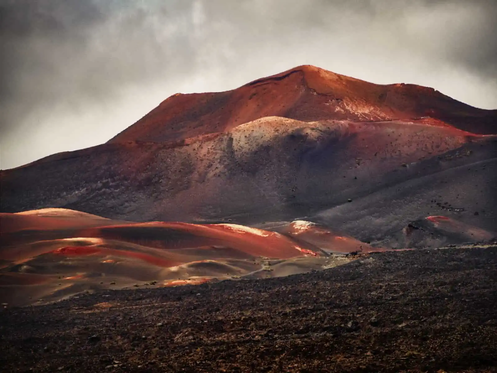 Image description: A landscape image which pictures a straight on shot of a volcano and its surrounding landscape. This image has browns, reds and yellows all throughout it showing soft curves of the volcano.