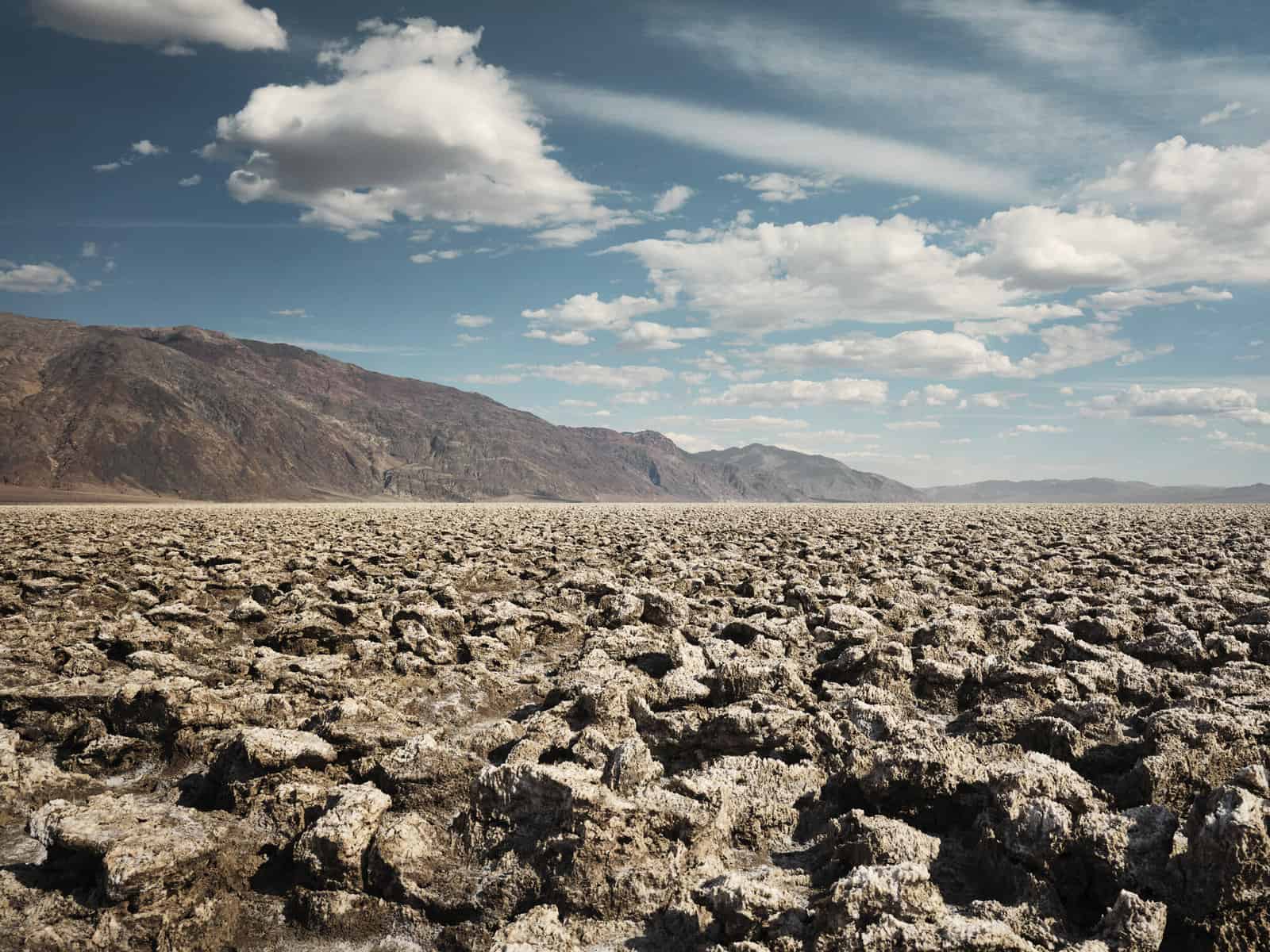 The image from Death Valley that inspired our further adventure planning. ID: A landscape image. In the foreground are rocks which are brown and sandy. In the background are desert mountains in reddy browns. The sky is blue with a few white clouds.
