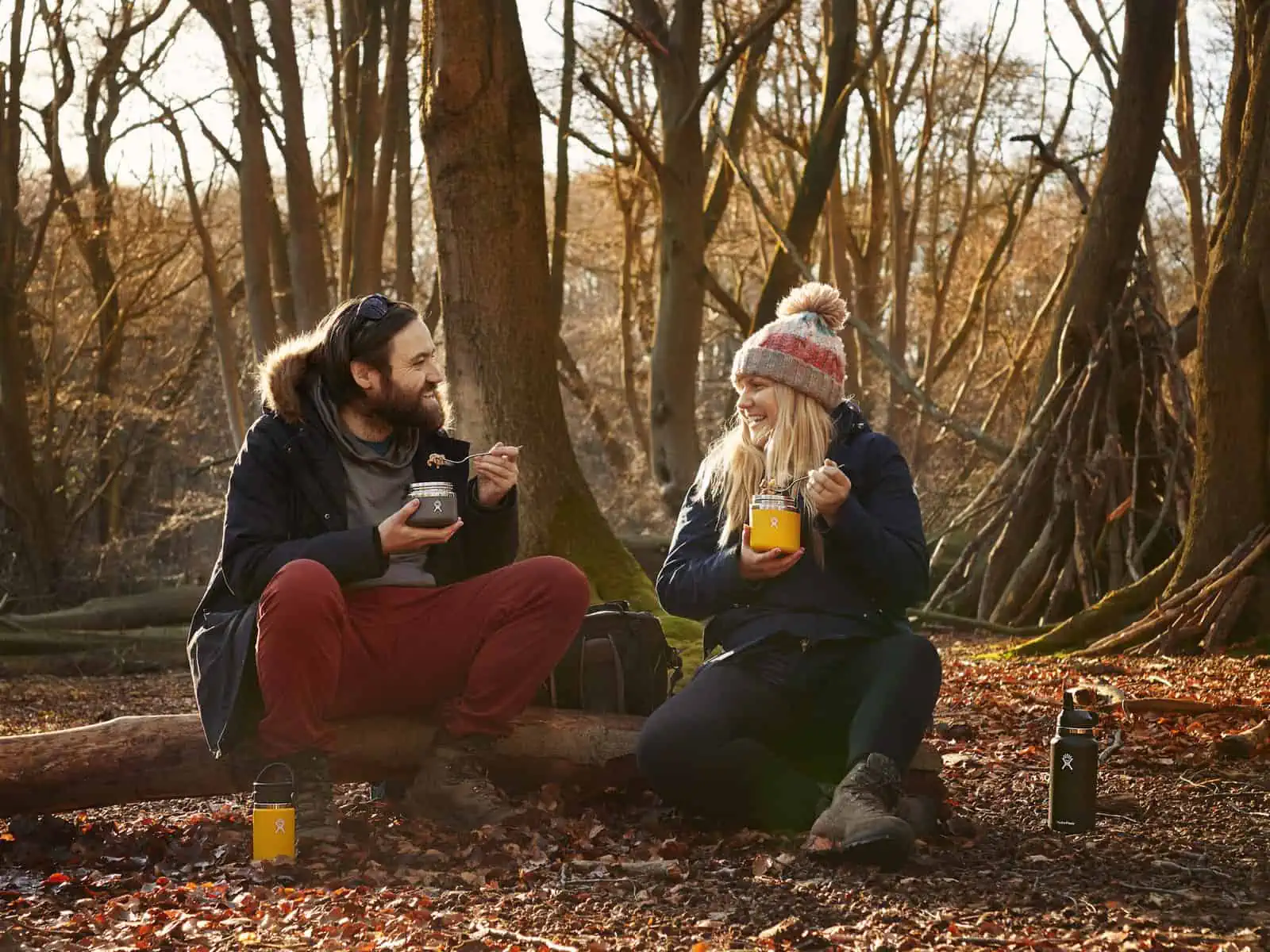Image Description: A landscape image. Fay and Matt sit in a forest on a log eating from Hydro Flask food containers. To Fay’s left is a black Hydro Flask. To Matt’s right is a yellow Hydro Flask. The ground is golden and covered with leaves. They ar…