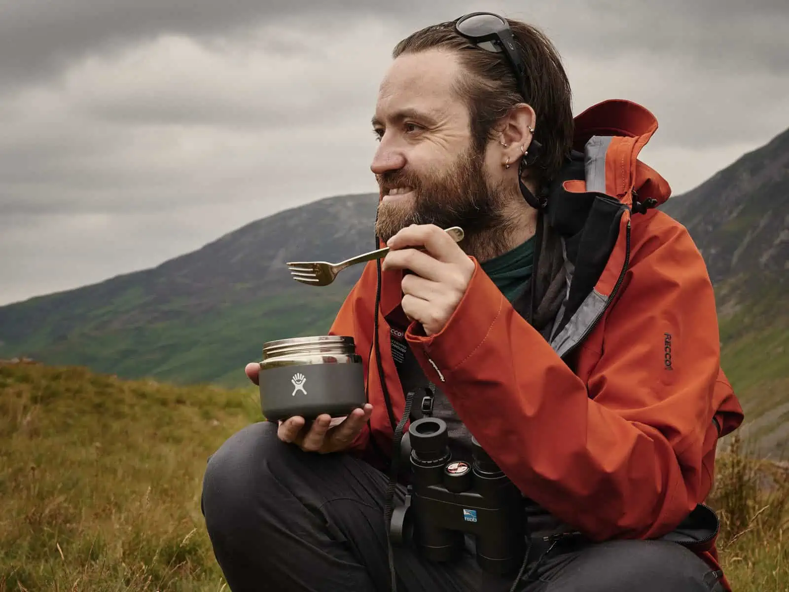 ID: A landscape image. Matt is sat on a rock eating from a food container with a fork. He is looking out at the view to his right and is smiling. He is wearing grey trousers, red coat and sunglasses on his head.
