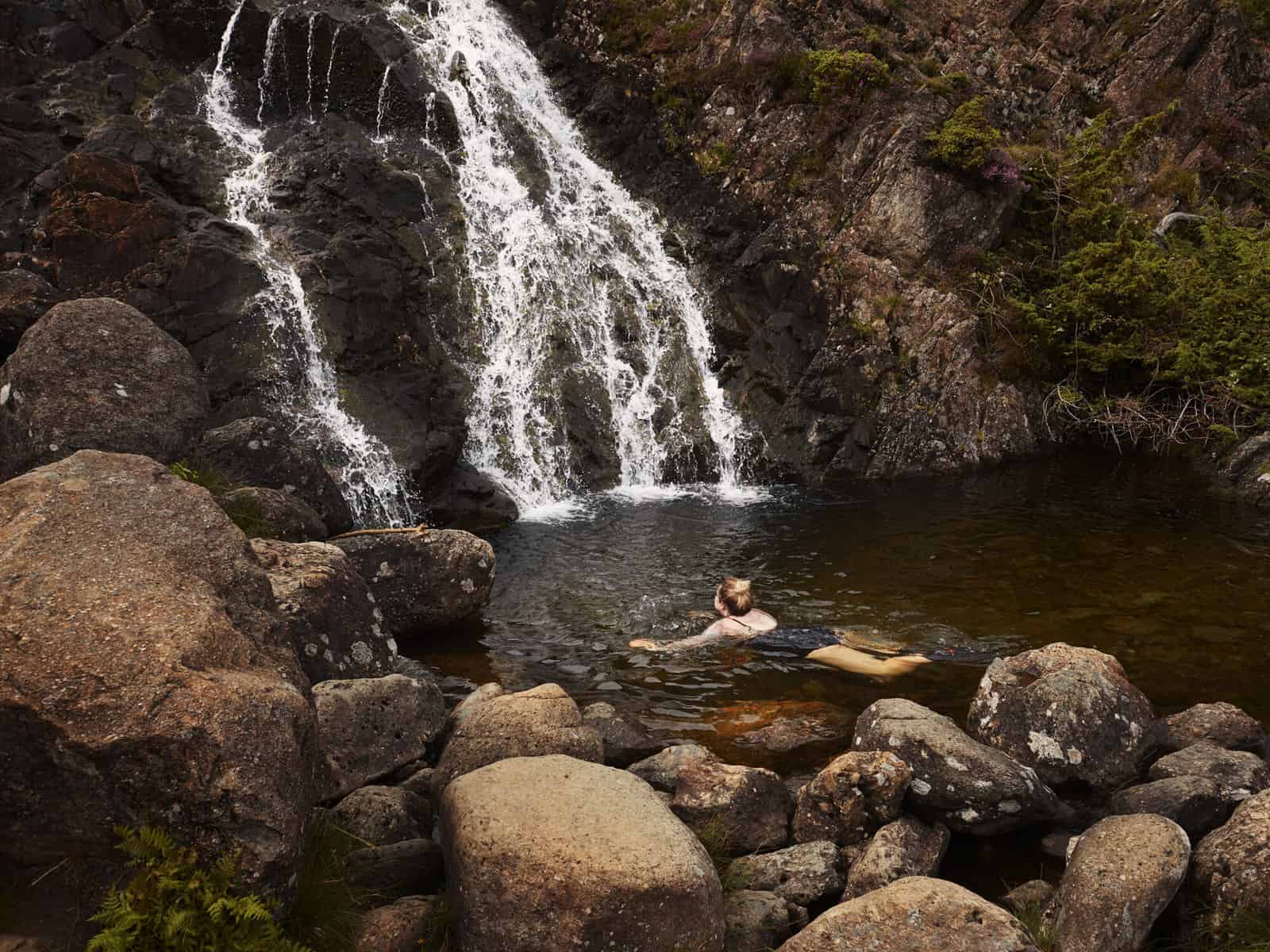 ID: A landscape image. Fay is pictured swimming in a wild swimming spot at the base of a waterfall. The water from above is white and fast flowing. Around the pool are lots of big rocks. The image is quite brown and Fay is looking at the waterfall wearing a black swimsuit.