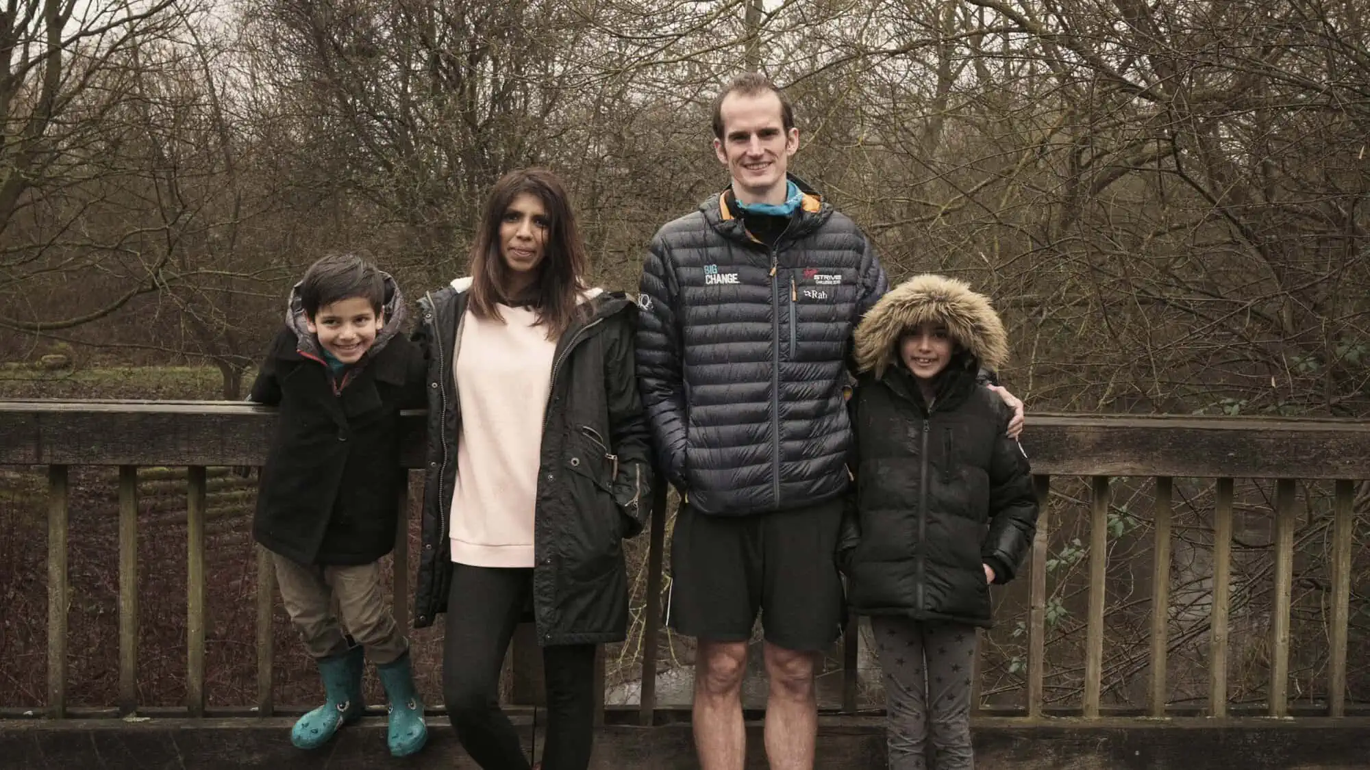 Image Description: A landscape format image. The Burton family stand on a bridge in front of an out of focus background of trees.
