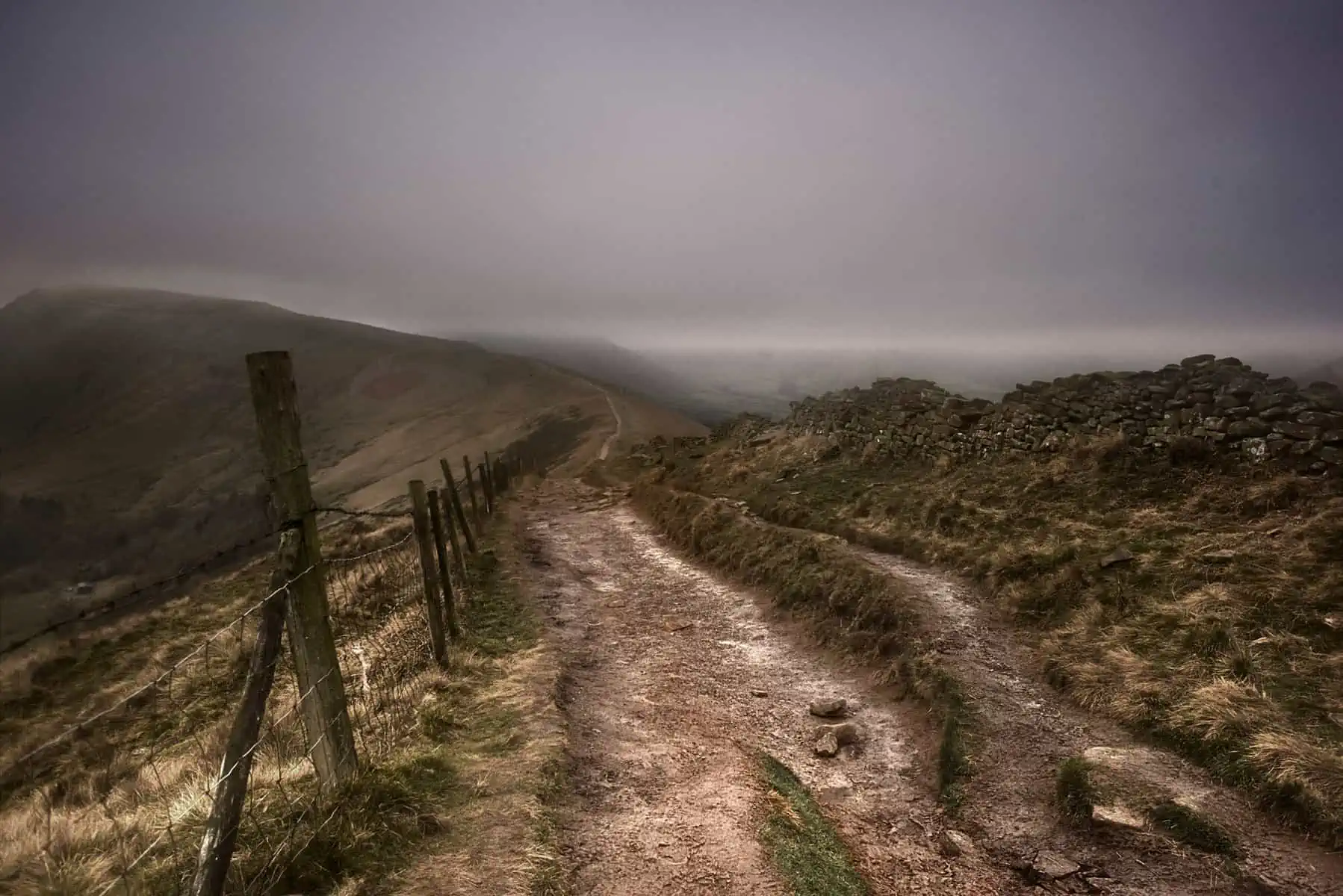 ID: A landscape image. A dark scene shows a dark and stormy day which looks very dramatic. In the foreground is a marked dirt path which leads to a hill in the distance. There are rocks on the right and a fence on the left. We cannot make out the back of the image due to cloud cover which is thick, heavy and grey.