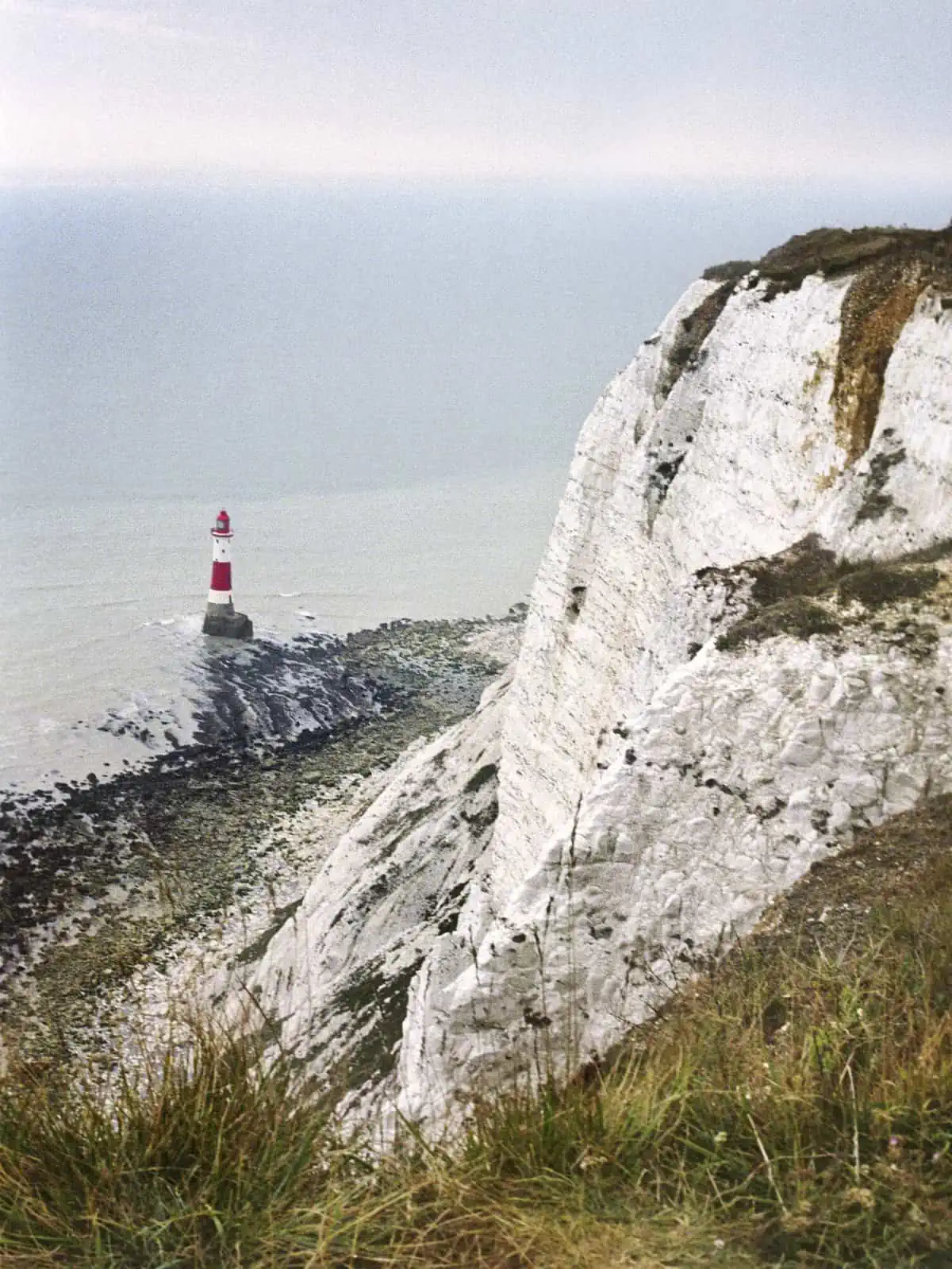 Image description: A portrait image. The white chalky cliff frames the right hand side of the image, and leads the eye down to the blue of the sea at the top left corner of the image. In the sea is a white and red striped lighthouse.