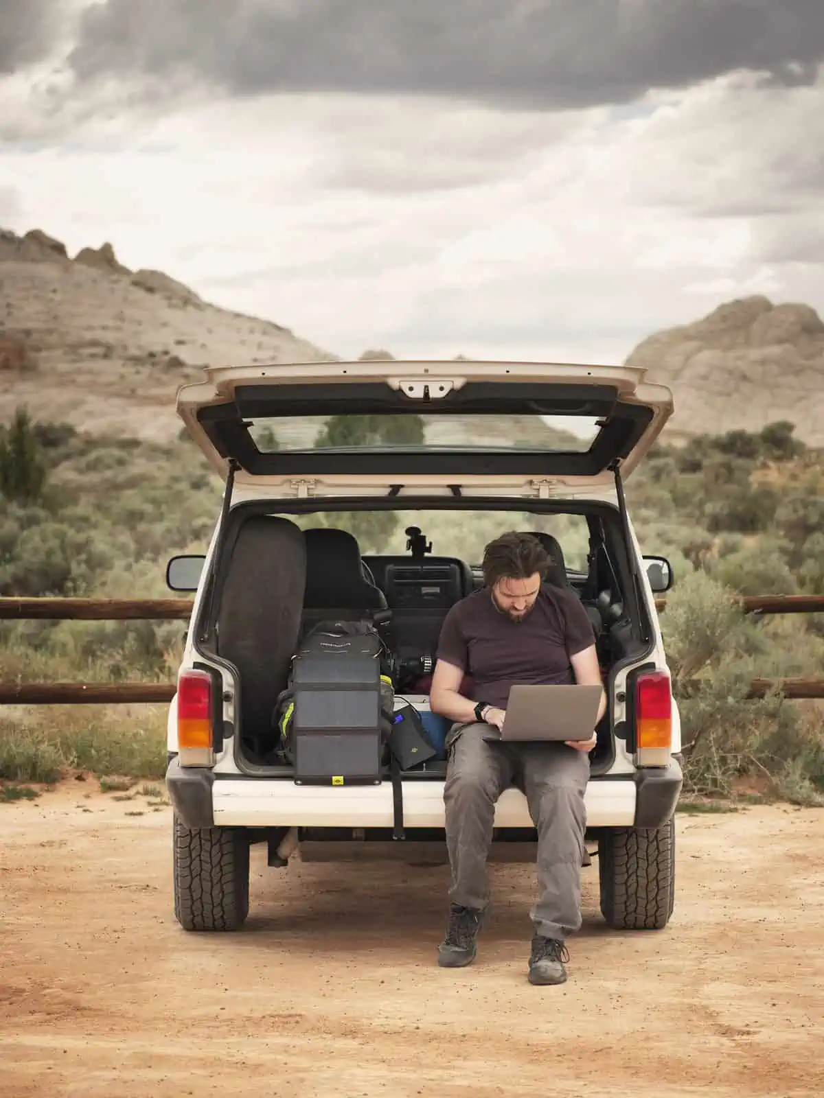 ID: A portrait image. Matt sits on the bumper of the boot of a 4x4 car. He is wearing grey trousers and purple top. He has a laptop on his knee and is working. To the side of him is a solar panel which is charging, and there are other photography kit items in the boot. The car is white and it is parked in a sandy spot. There is a fence behind the car, in the background are mountains.