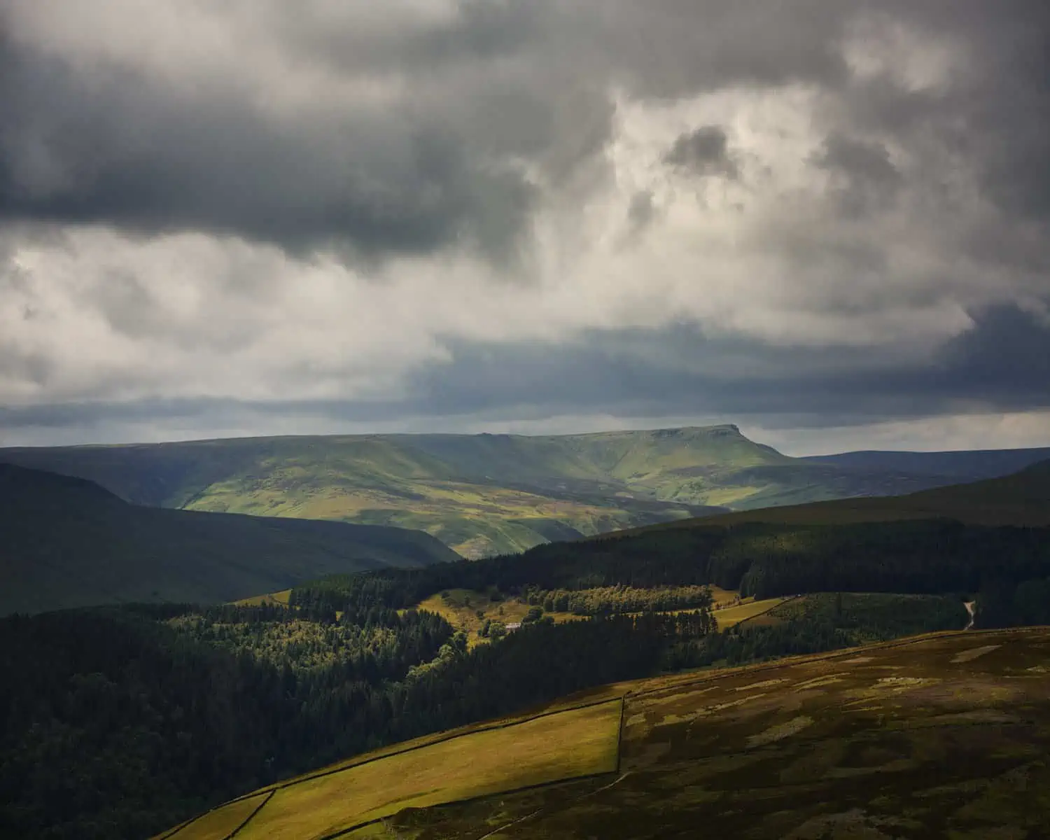 ID: A landscape image. A dark, moody scene with dramatic shadows from clouds over the moorlands. The fields and hills almost look like velvet. We see greens, browns and blues in this scene with geometric hill shapes in the background.