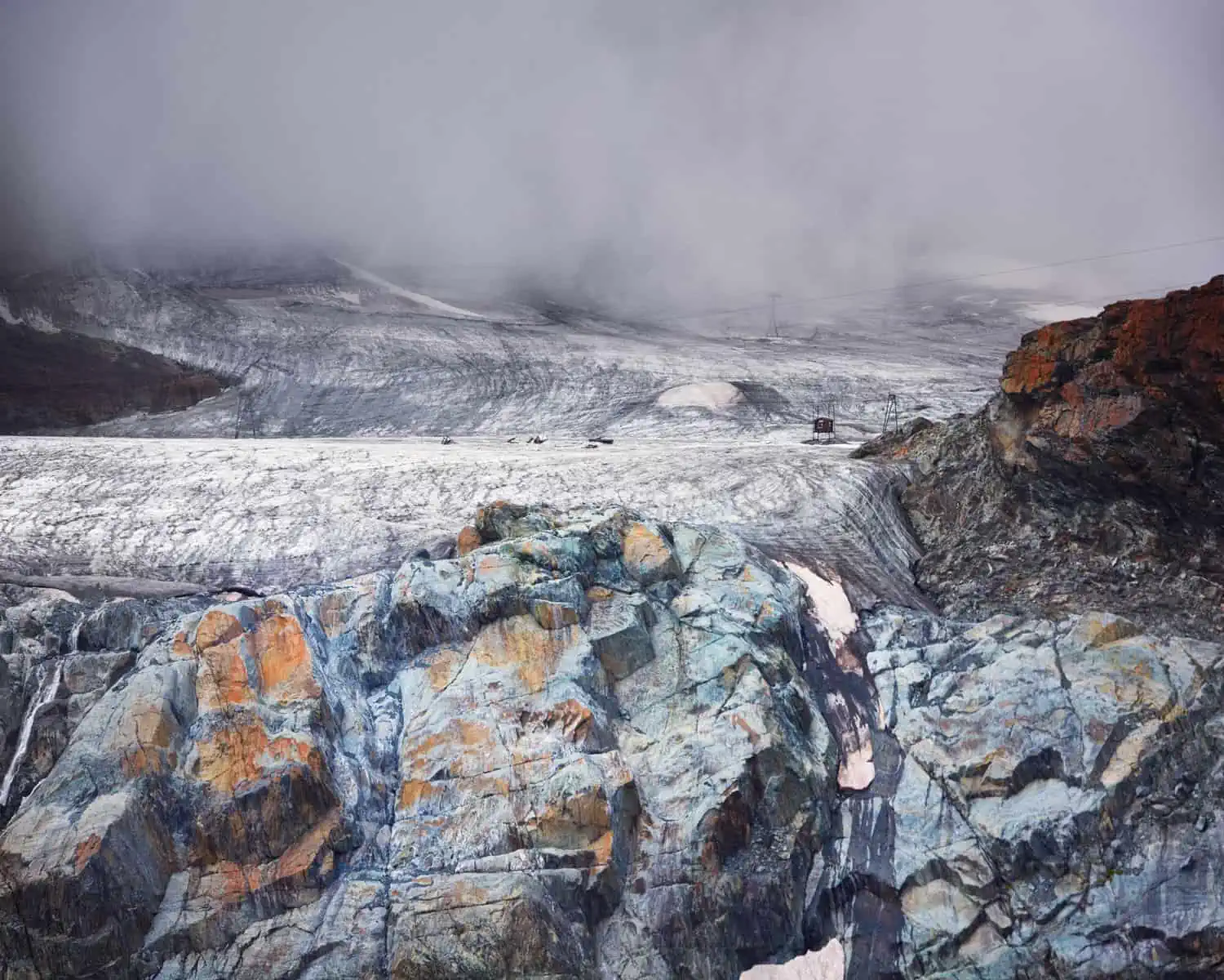 Image Description: A landscape format image. A glacier is in the foreground. It is mostly blue ice, but there are also yellow patches of lichen. In the background we see an ice filed, hardware for ski lifts and thick clouds at the top of the frame.