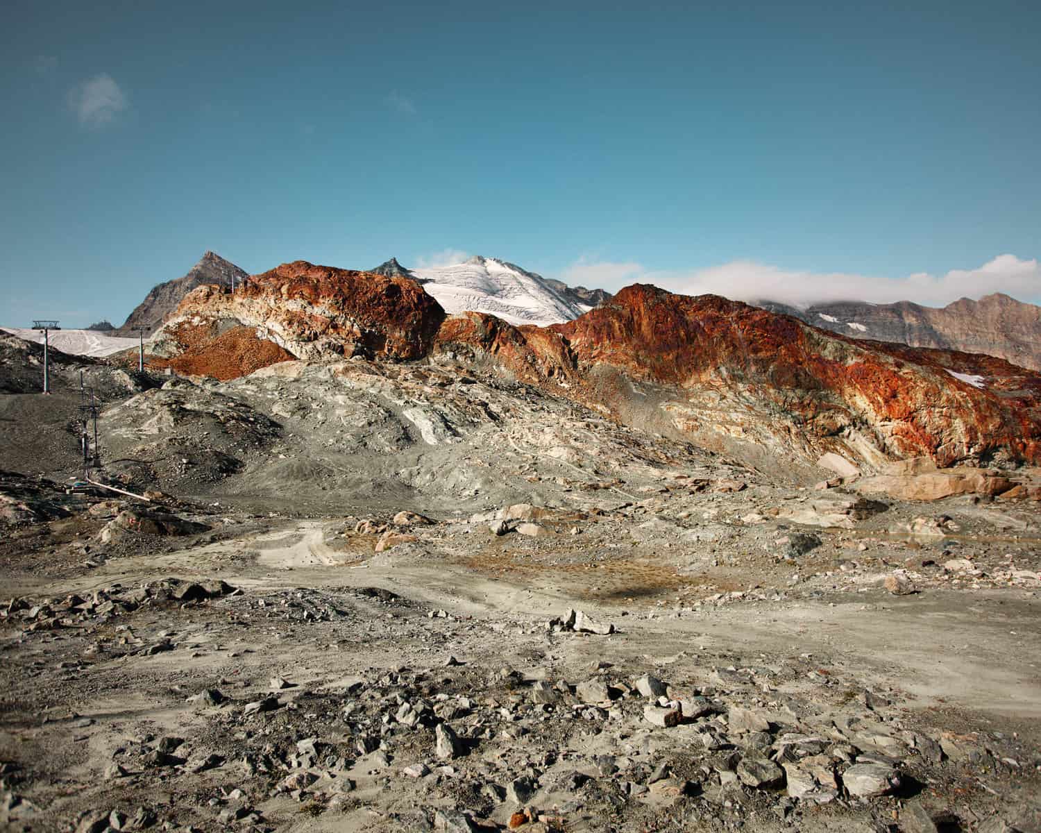 Image Description: A landscape format image. A grey-yellow almost desert landscape dominates the foreground. There is a clear blue sky above and striking orange rock in the hills in the background. The image looks like it could have been made somewhere very hot, except for the fact that there is a glacier, snow-capped mountains and ski lift hardware visible in the far background.