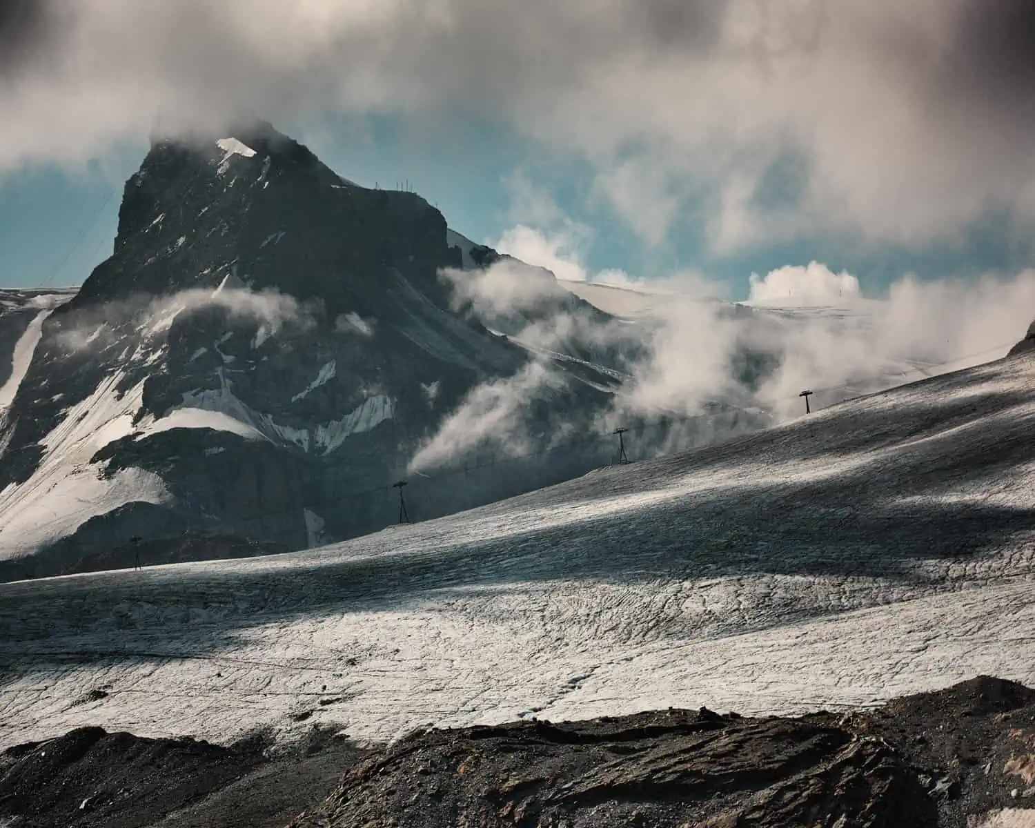 Image Description: A landscape format image. The image is dominated by the ice field of a glacier. We see shadows cast by low clouds which are also visible in the image whilst a high mountain peak towers in the background. A ski lift runs diagonally through the frame. A band of dark rock dominates the foreground. The colours are cold and neutral.