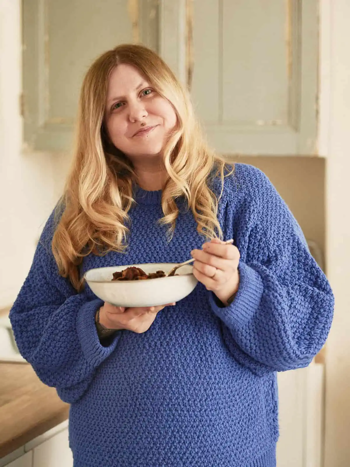 Image Descriptions: Portrait format image.&nbsp;&nbsp;Fay stands in the centre of the frame holding a bowl of food and fork.&nbsp;&nbsp;Fay wear an oversized blue jumper and there are cupboards and a counter top in the background.&nbsp;&nbsp;