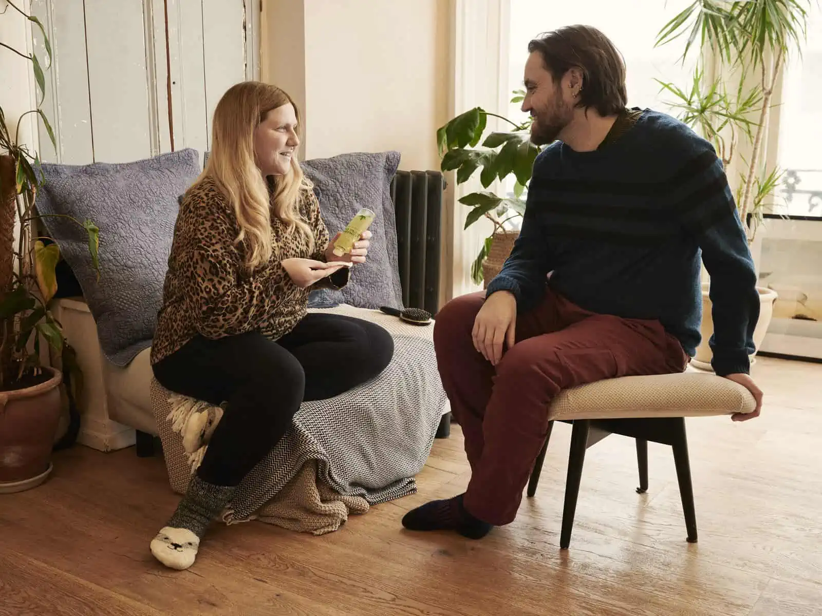 Image Descrition: a landscape format image.&nbsp;&nbsp;Fay and Matt sit on a chair and small stool in a bright, airy room, surrounded by plants.&nbsp;&nbsp;Fay is wearing a leopard print top, black leggings and woolly socks and is using an Avon prod…