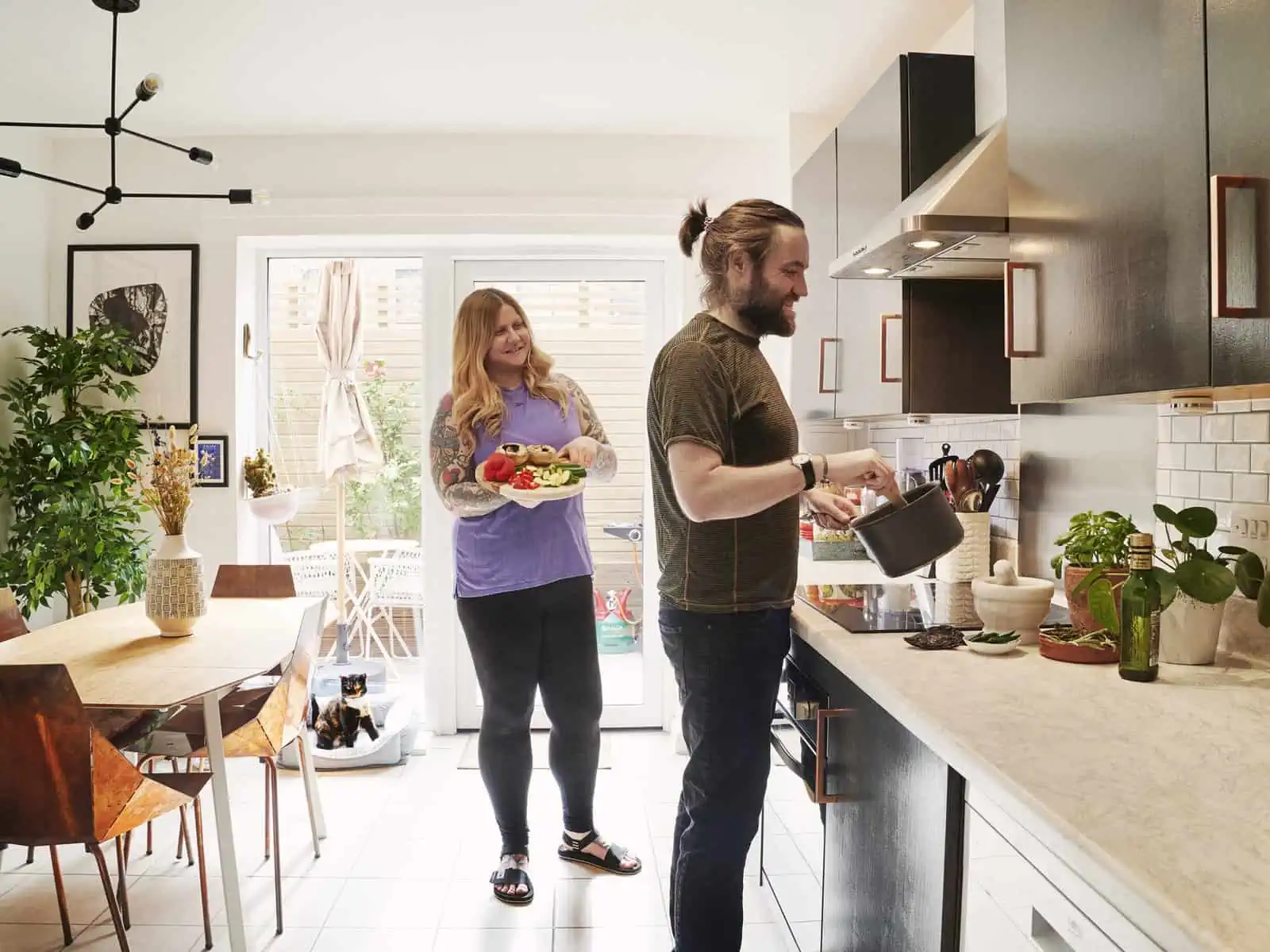ID: A landscape image. Fay and Matt are cooking together in their kitchen. It is bright and airy and decorated in a modern way. Matt stands by the stove and is using a spoon in a pan whilst Fay brings toward him a tray of vegetables ready to be cook…