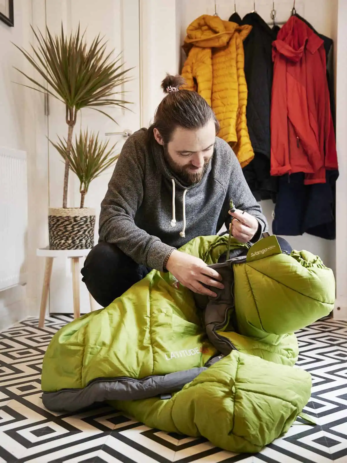 ID: A portrait image. Matt is crouched on the floor checking a green sleeping bag and prepping equipment. He is wearing a grey hoody. The floor is a black and white pattern, with plant in the background and some coats hung up behind. It is a light …