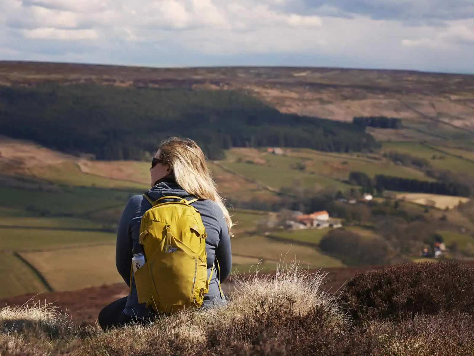 Image description: A landscape image. Fay sits at the top of the hill to the left of the frame and is facing away from the camera. Hair is over the right shoulder and Fay looks out to the left with black sunglasses on. Fay wears a yellow bag with a blue jacket and black trousers. There is a silver bottle in the side pouch of Fay’s bag. Fay takes in the view of the hills in the background which have green fields along them. The sky is blue with some clouds.