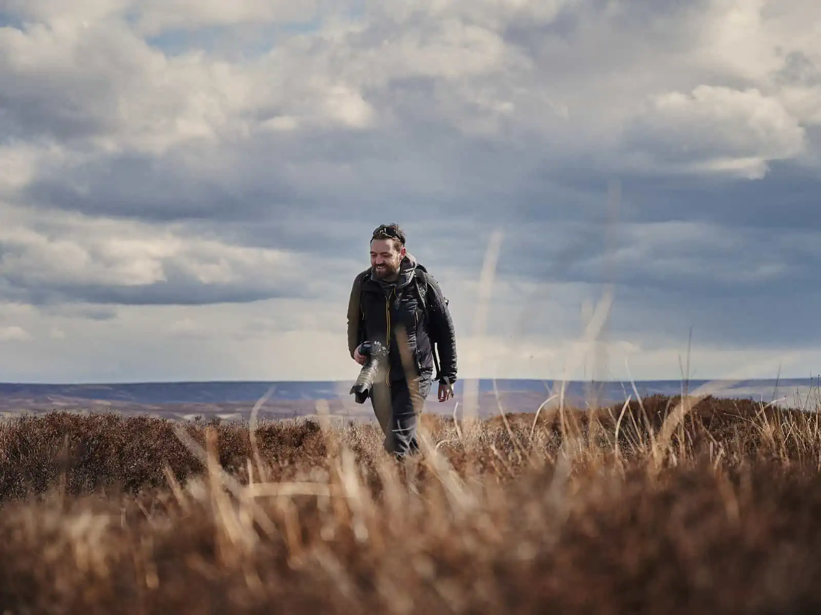 Image description: A landscape image. Matt is in the centre of the frame and is walking forward with the gaze slightly to the left side. Grasses in the bottom third of the frame are obfuscating his legs, and these are brown and yellow in colour. Matt wears a mostly black outfit and is carrying a camera with a long lens. In the background there are hills and a dramatic blue sky takes over more than half of the frame.