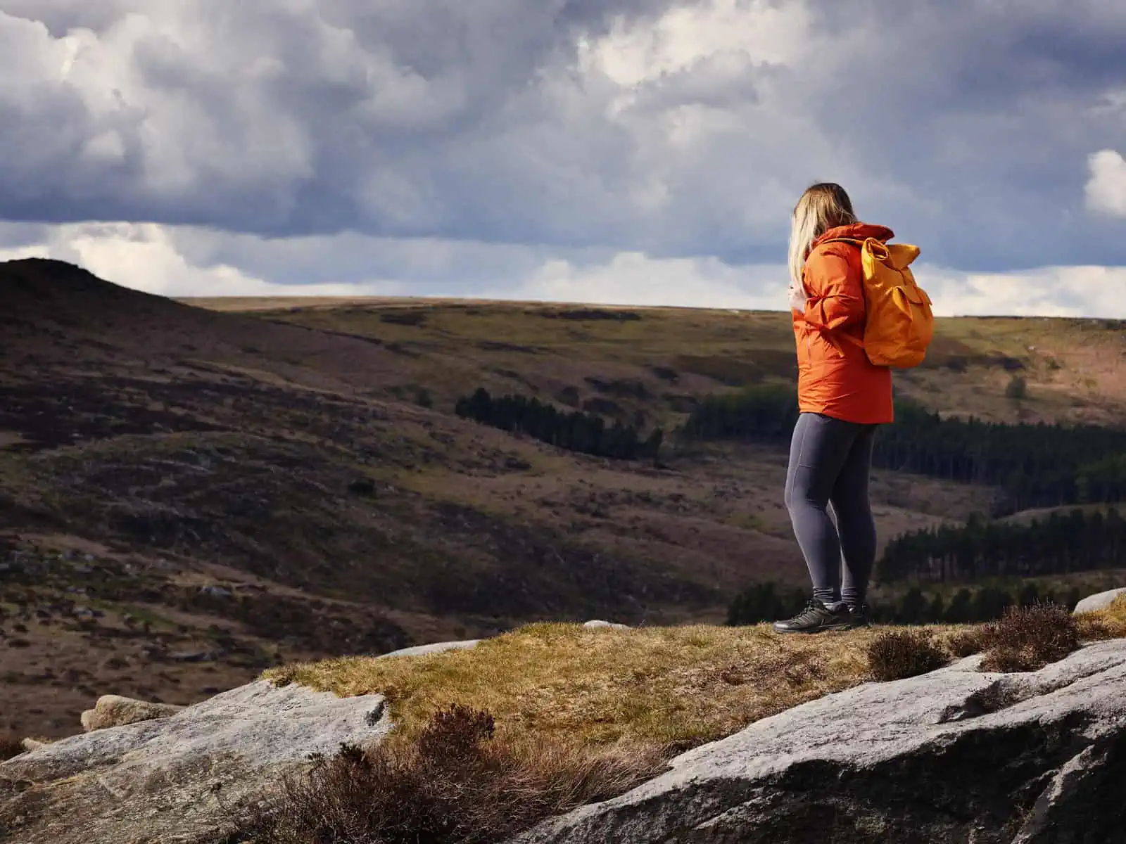 ID: A landscape image. Fay stands to right of the frame and is turned to the side in side profile. Fay wears grey trousers, black shoes, bright red coat and yellow bag. Fay stands on a section of mossy hill with rocks formed in the hills around. In the background are large, undulating hills which are green and brown. The sky is quite dark with sections of open blue sky which are casting a dramatic shadow onto the hills around and casting a spot light on Fay.