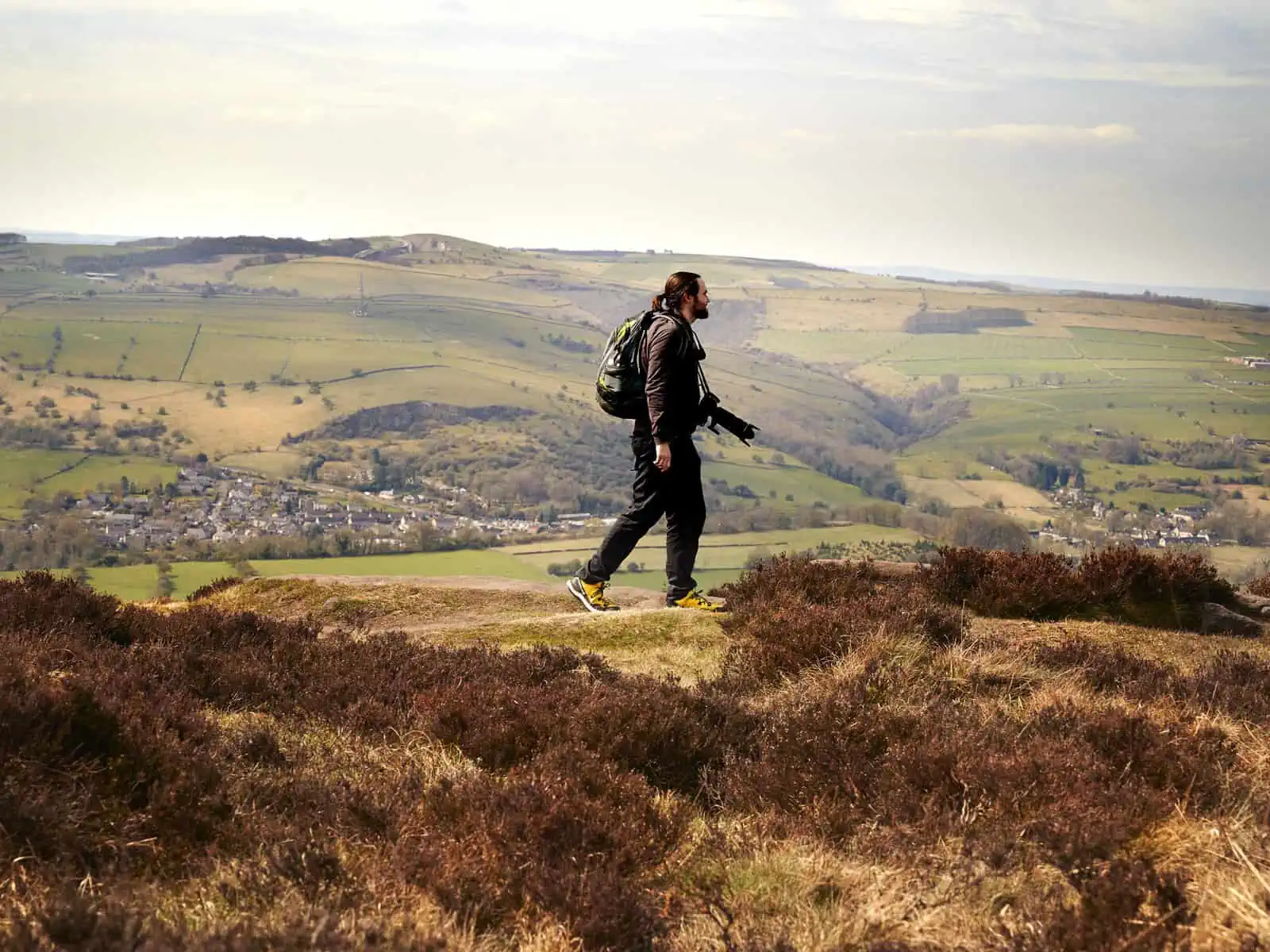ID: A landscape image. Matt walks from left to right across the top of a hill. In the foreground is mossy grass and rocky crops. Behind him are hills which are greens and browns. The sky is a pale blue. He wears yellow shoes, black trousers and grey hoody. His bag is blue and green and he has a black camera with long lens around his neck. It looks to be a warm sunny day.