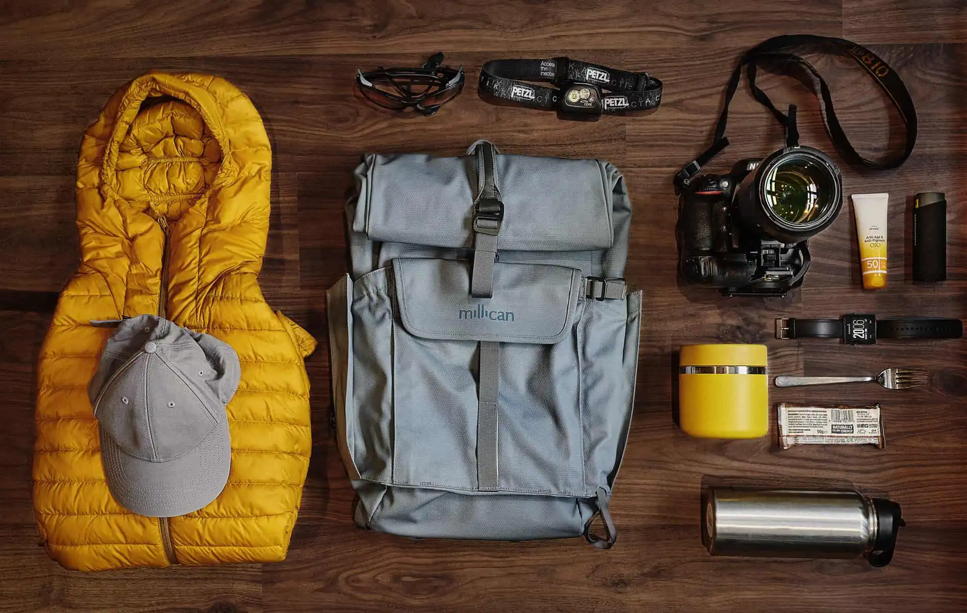 Image description: A landscape image. A flat lay on a dark wood floor shows the items kept in a day pack for exploring. A Millican ‘The Mavericks’ Smith the Roll 15l pack in Tarn sits in the centre. To the left is a bright yellow puffy jacket with grey baseball cap on top. Above the pack is a pair of black sunglasses and a black head torch. To the right is a camera, a tube of yellow sun cream and a battery charger. Below, a yellow food jar, a black smart watch, a silver form, an energy bar and at the bottom a titanium water bottle.&nbsp;