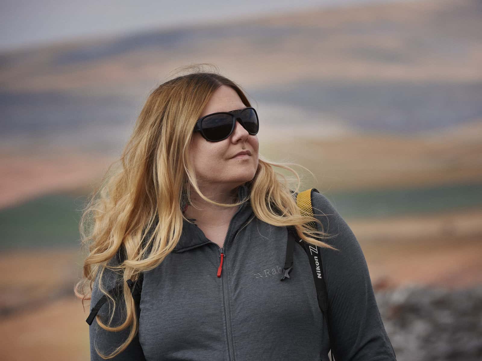 ID: A landscape image. A close up of Fay with head turned to the right. Fay has hair down, is wearing a blur zipped up jacket, yellow camera strap is on the right hand side and is wearing black sunglasses. Fay smiles. In the background is blown out …