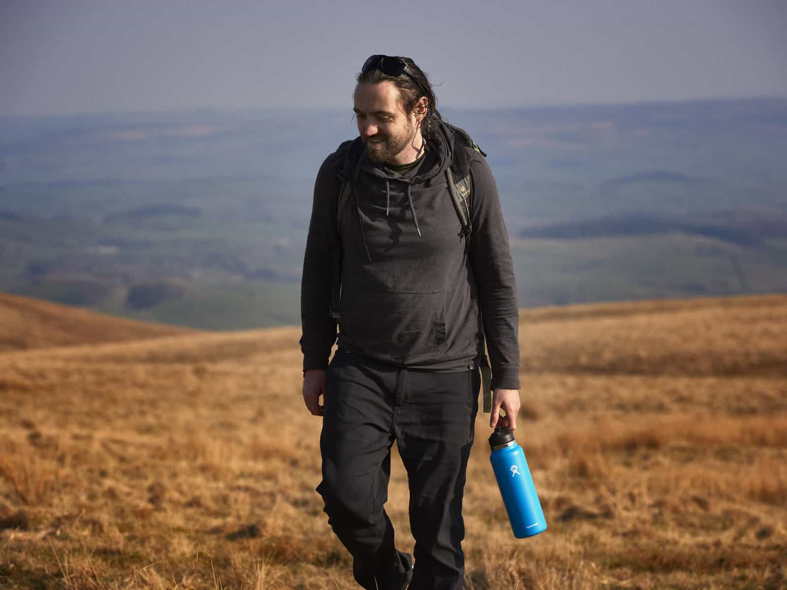 ID: A landscape image. Matt is in the foreground and central in the picture. He has his head turned to one side and is smiling and holding a blue reusable water bottle. He is wearing black trousers, grey hoody and a grey strap of a backpack can be s…