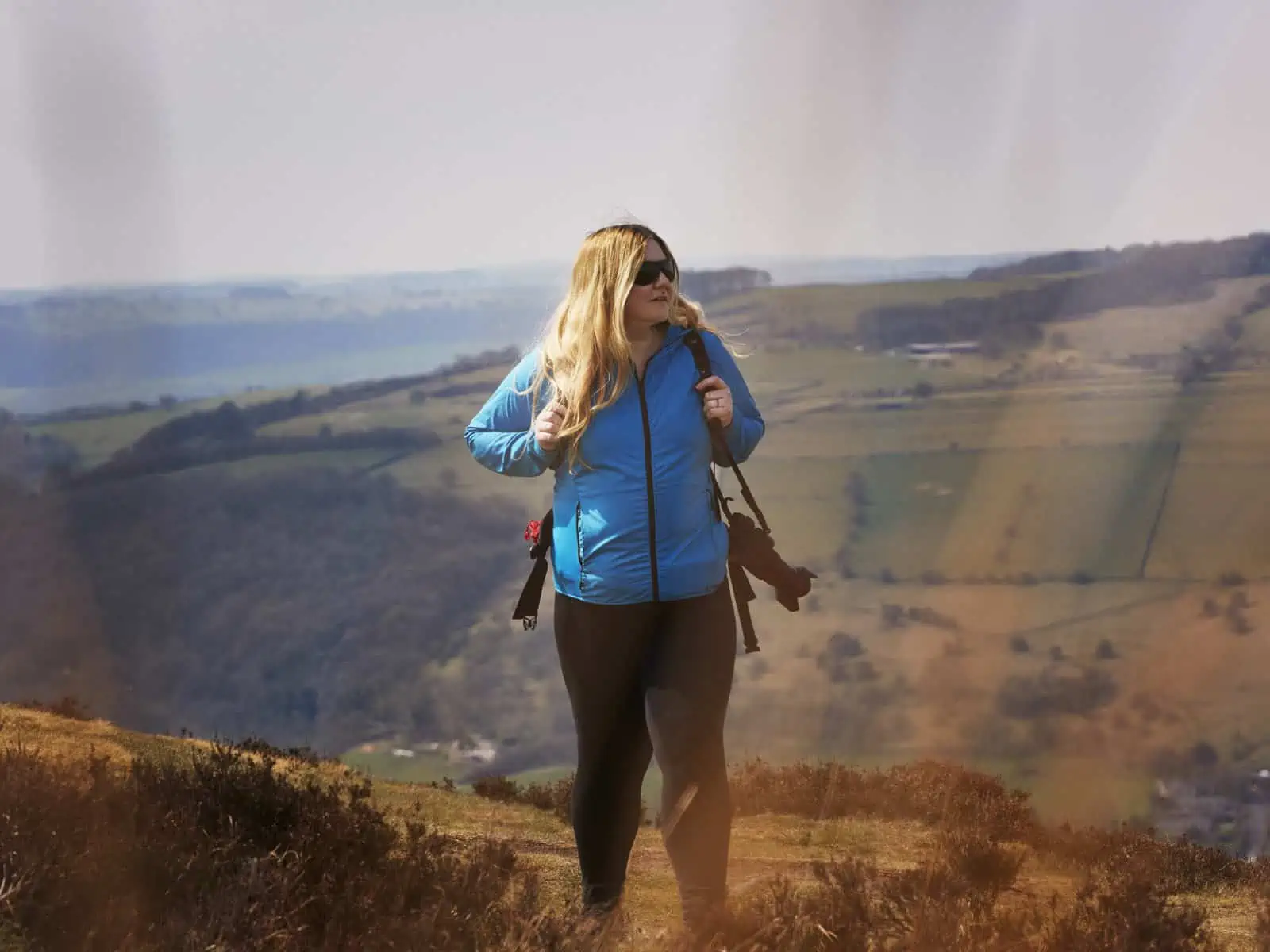ID: A landscape image. Fay is walking centre of the frame up a hillside. In the foreground are out of focus hazy grasses. In the background are steep hills. The ground is green and brown. Fay is wearing black trousers, blue jacket and black sunglasses. A black camera is over Fay’s shoulder.