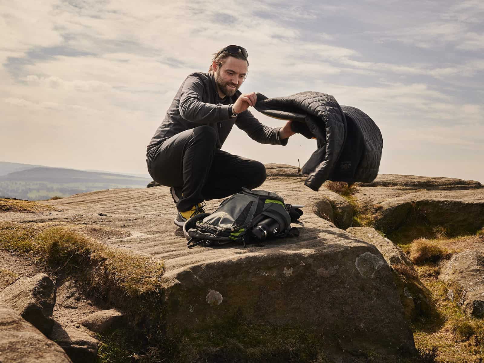 ID: A landscape image. Matt sits on a rocky outcrop taking his coat out of his backpack and opening it out. Whilst he does this, a gust of wind has blown the edges away. He is crouched down over his backpack which is grey and green. He is wearing ye…