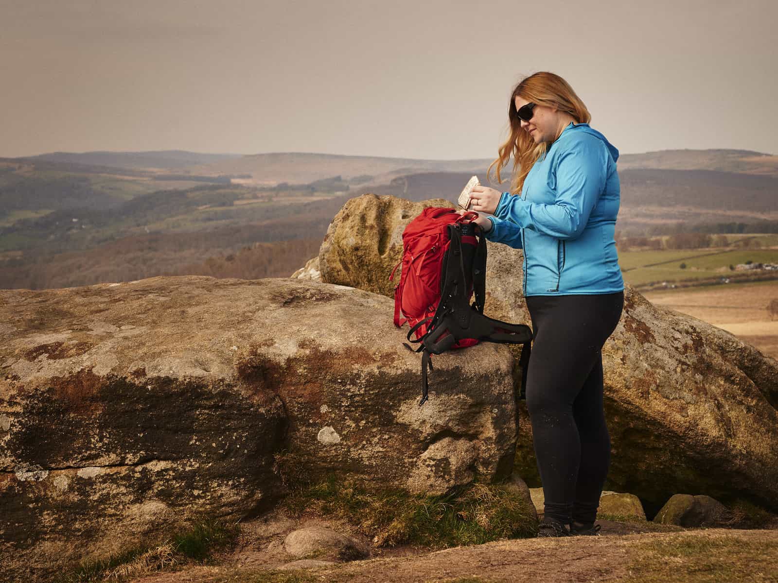 ID: A landscape image. Fay stands to camera right and is wearing black trousers, blue jacket and black sunglasses. Fay’s bag is perched on a rock and the red backpack is open. Fay is putting an empty food wrapper into the top of the bag. The rocks a…
