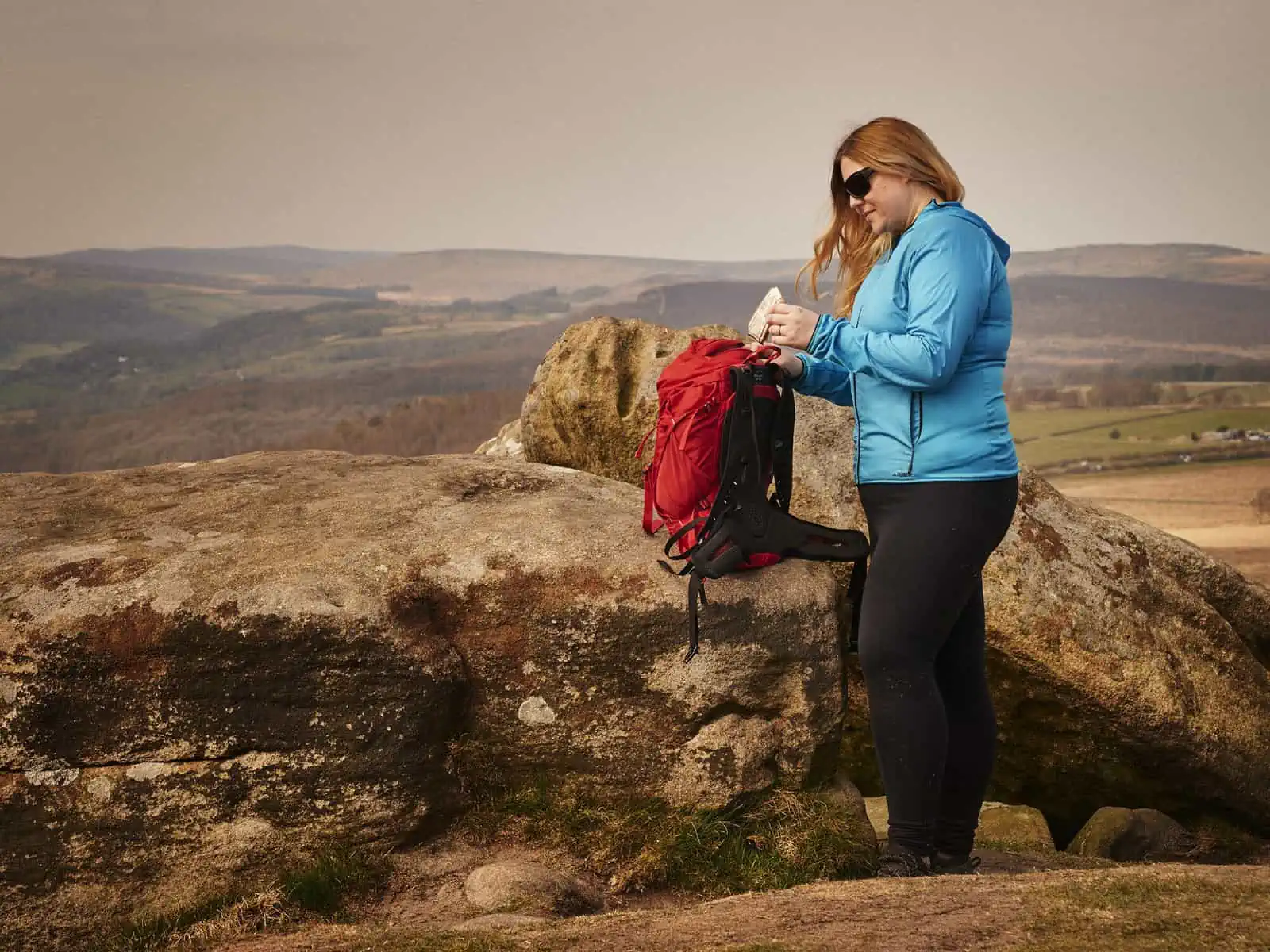 ID: A landscape image. Fay stands to camera right and is wearing black trousers, blue jacket and black sunglasses. Fay’s bag is perched on a rock and the red backpack is open. Fay is putting an empty food wrapper into the top of the bag. The rocks a…