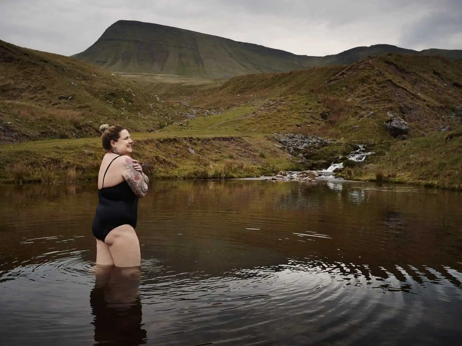 ID: A landscape format image. Fay is wearing a swim suit and is stood in a mountain pool having just been wild swimming. There is a waterfall and a mountain in the background.