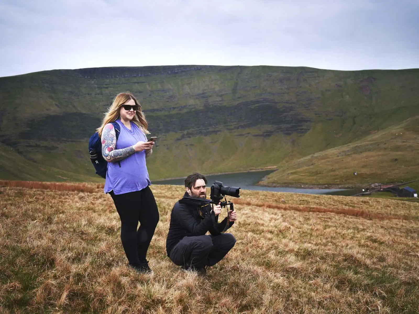 ID: Fay and Matt are close to each other in the centre of this landscape image. Fay stands to the left, wearing purple top and black trousers with blue bag. Fay holds a phone and is logging shots as they shoot a video. Matt is crouched down, wearing a black coat, black trousers and is holding a camera on a shoulder rig. They are stood on the top of a hill, which is brown and grassy. In the background are higher, green mountains and a lake underneath.