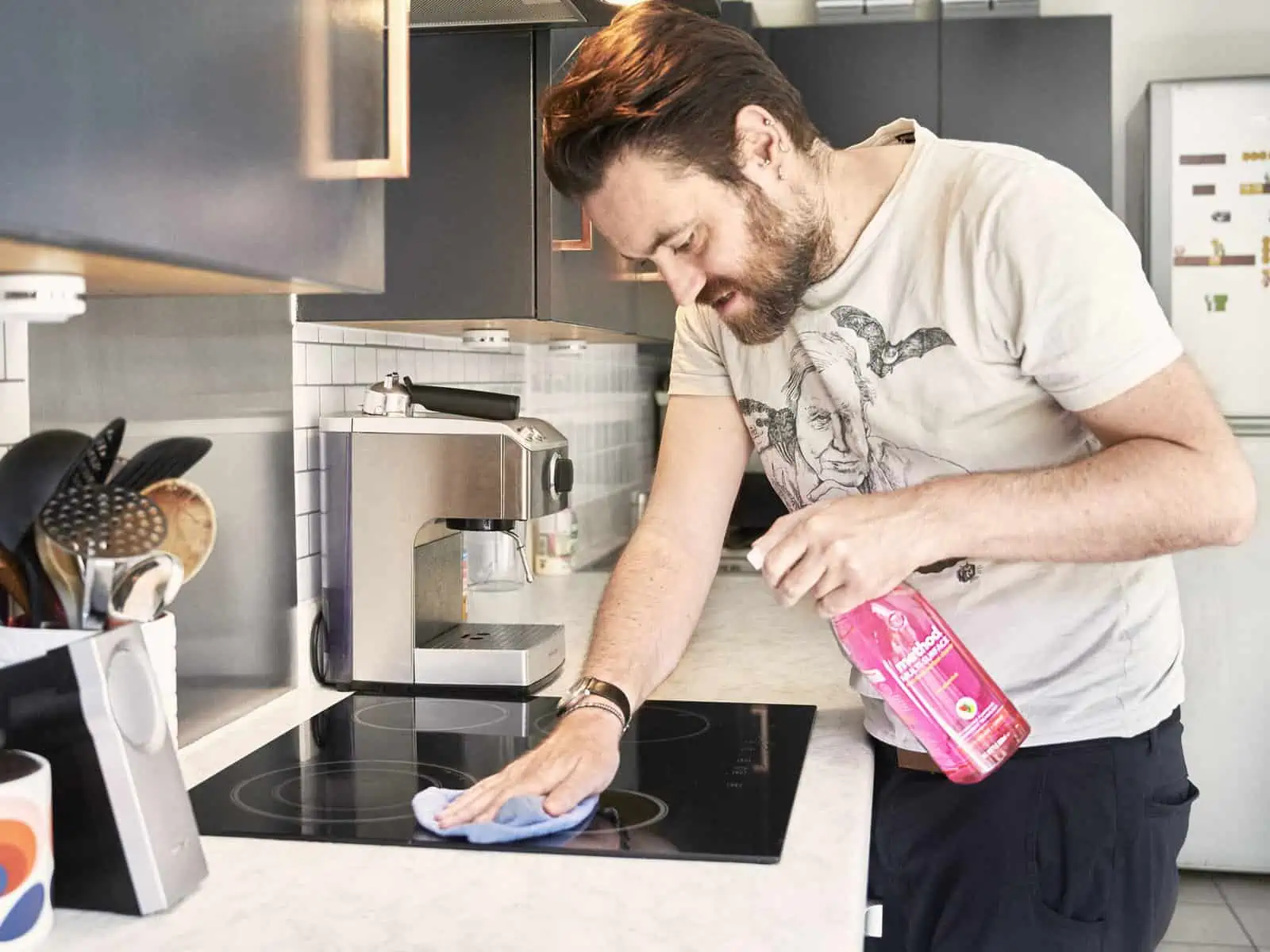ID: A landscape image. Matt cleans cooker in the kitchen and is using Method cleaning spray. We love Method as their claims are easy to find and back up about their sustainable products. The product is pink, he holds a blue both and is wearing a whi…