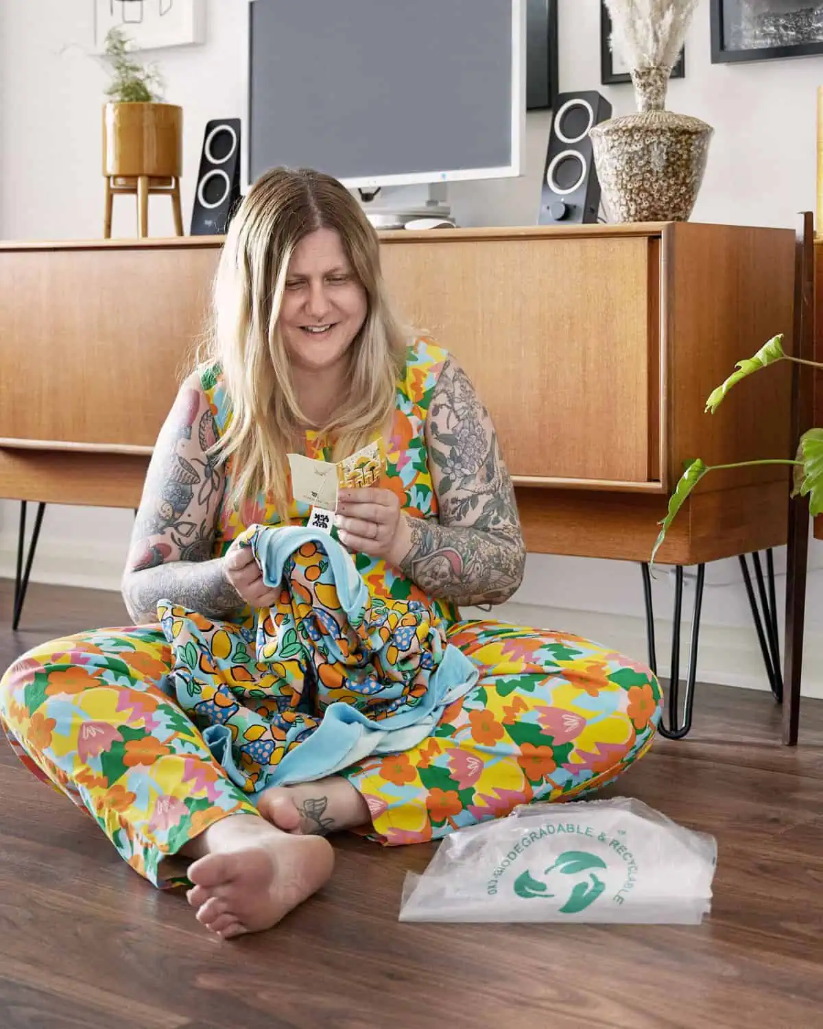 ID: A portrait image. Fay sits on the floor, behind Fay is a mid century furniture, with computer on top and pictures on the wall. Fay is wearing a Lucy &amp; Yak boiler suit in bright pattern. In Fay’s hands are a Lucy and Yak jumper, and Fay is reading the label which goes into detail about their sustainable manufacturing process and biodegradable packaging.
