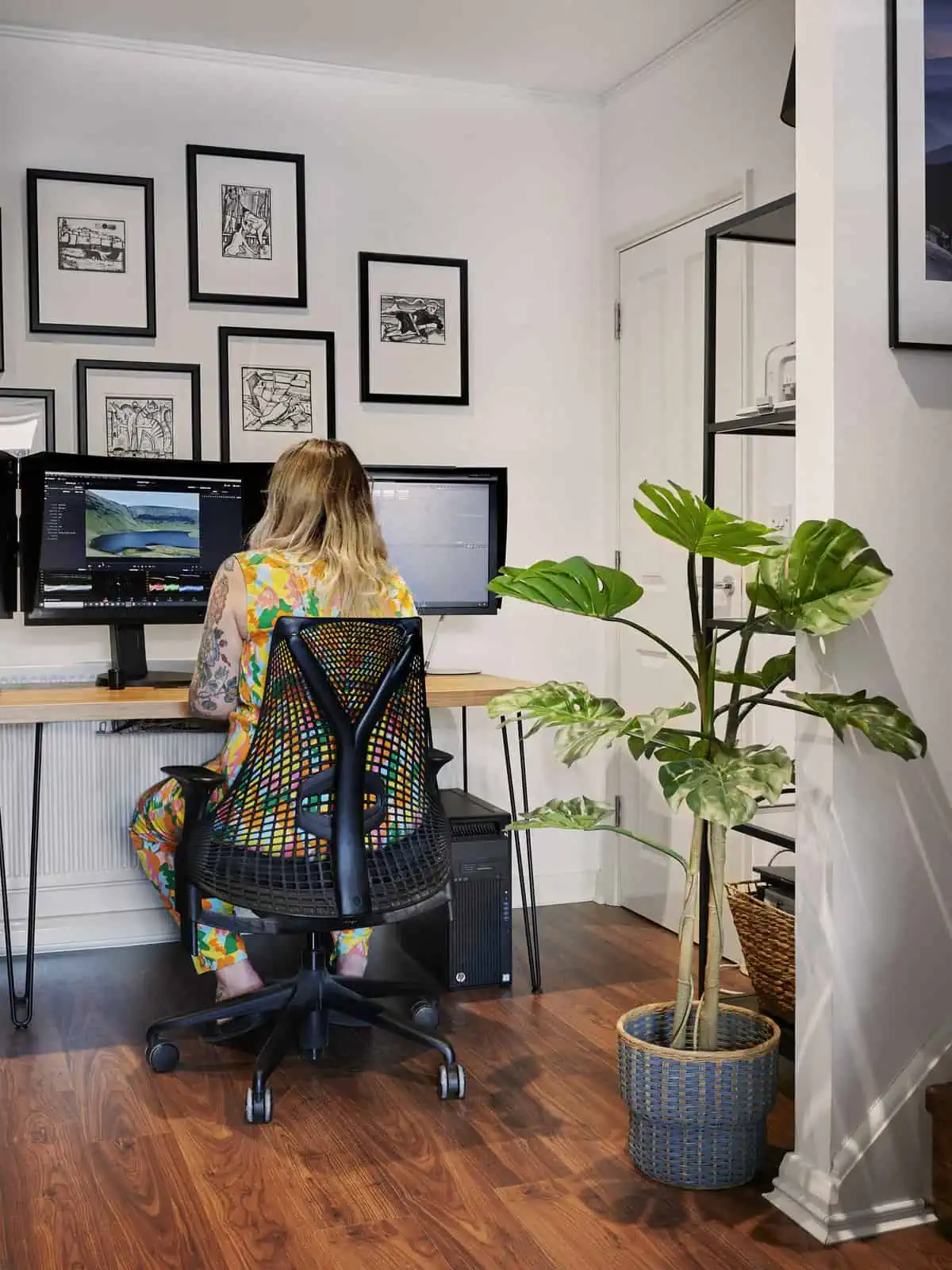 ID: A portrait image. Fay is sat at their desk in the studio. In the foreground is a plant, a wall and shelves. Sat at the desk, Fay is using their computer with two screens on a desk with hair pin legs. Fay is sat in a Sayl chair and is wearing a bright patterned jumpsuit. On the screens is editing software.