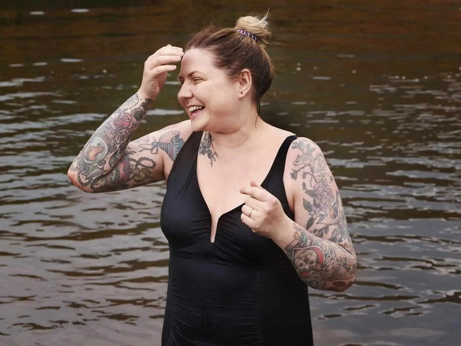 ID: A landscape image. Fay is captured from head to low torso standing in the water. Fay wears a black swimsuit and is laughing. Fays right hand is up to the face brushing water off from the swim.