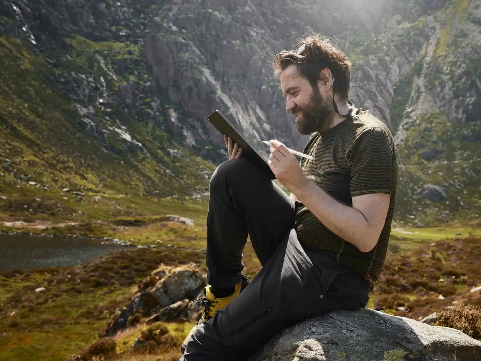 ID: A landscape image. Matt is sat on a rock in a mountainous, slightly out of focus scene working on his iPad and working up an image on Affinity Photo on location. He is wearing yellow shoes, black trousers, green top and is using an iPad with a white Apple Pencil.