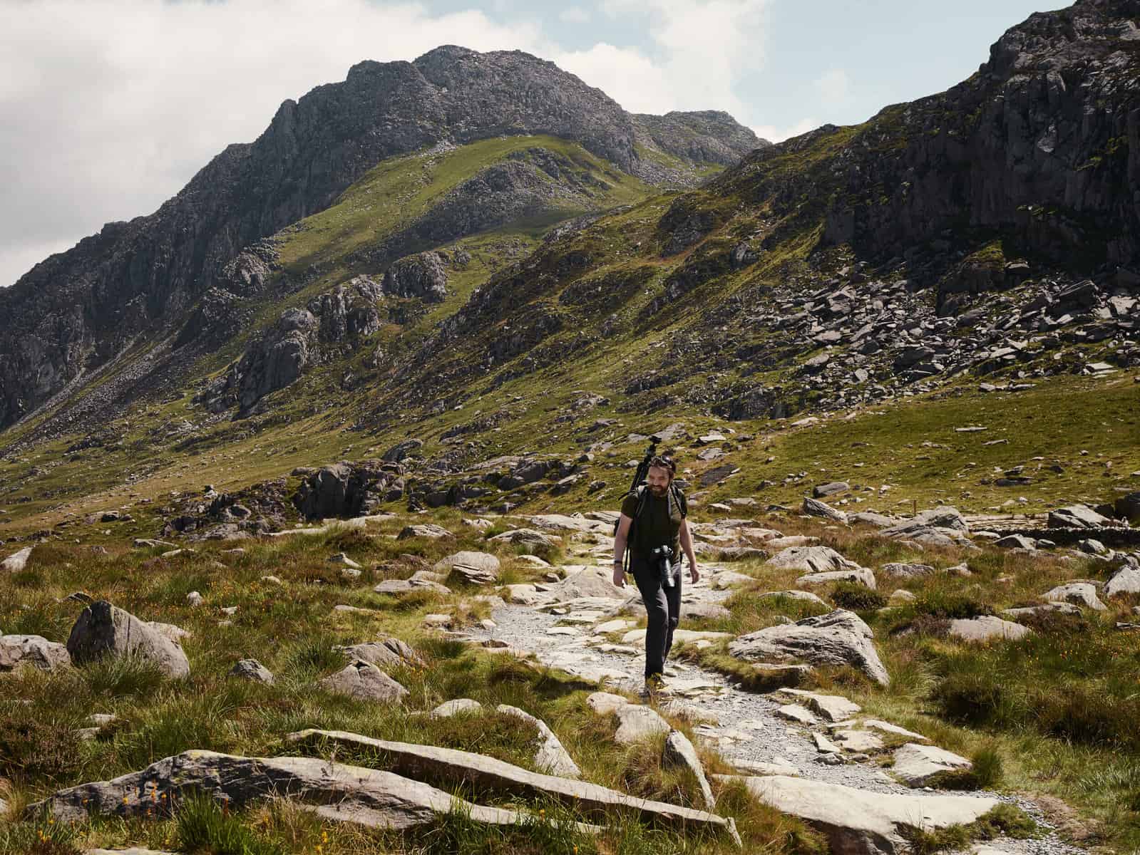 ID: A landscape image. Matt is walking to the front of the frame on a Rocky Mountain path with rocks and grasses all around him. He is wearing black trousers, yellow shoes, green top and is carrying a camera around his neck and a tripod is visible from his backpack. As the trail heads off behind him, we see rocky mountains above him that are slate grey. The sky is slightly overcast on the left side and is blue on the other.