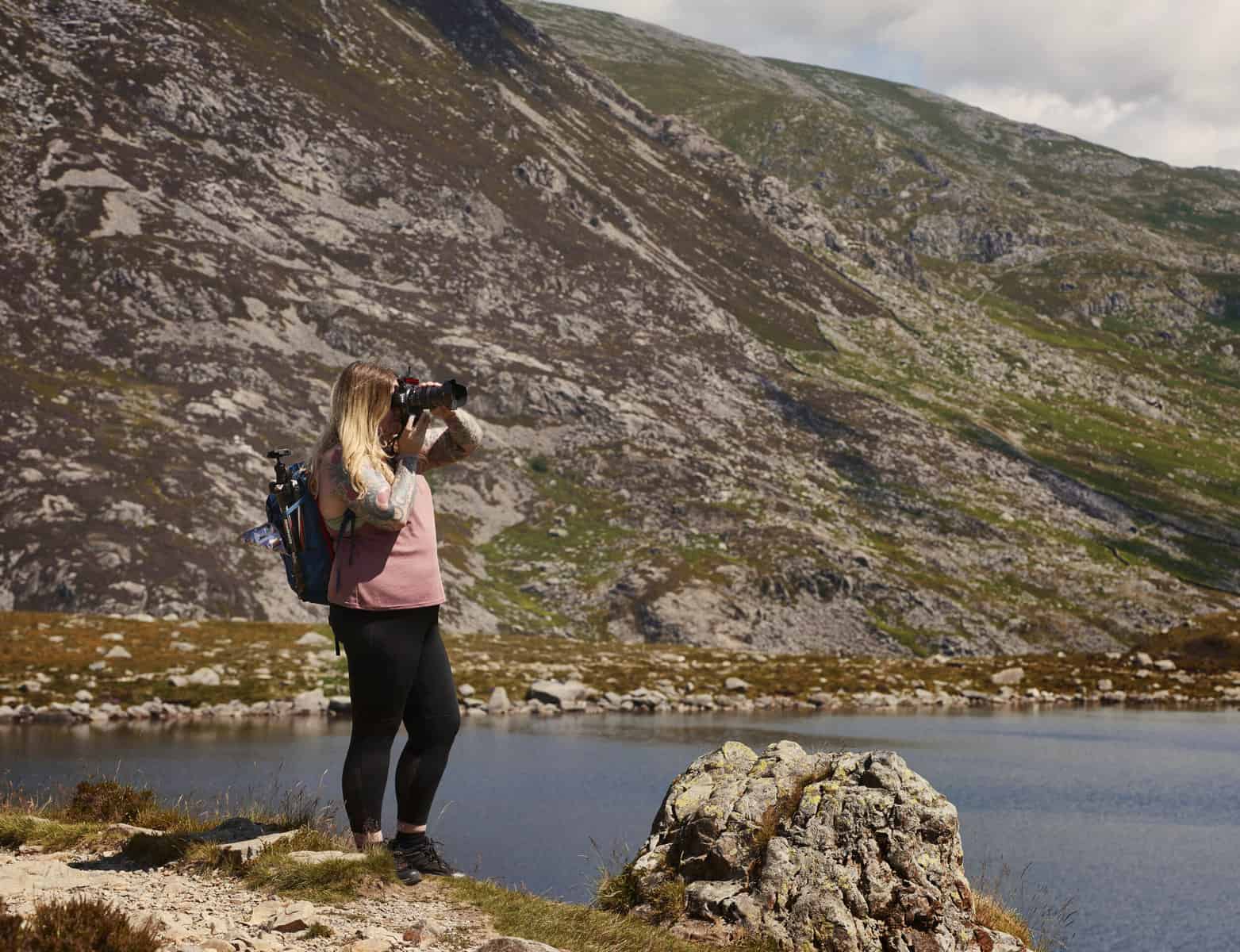 ID: A landscape image. Fay stands pictured full length on the left hand side of the frame with backpack holding a tripod. Fay is taking a picture handheld and wears black shoes, black trousers and a pink top. In the background is a lake and a rocky mountain scene out of focus.