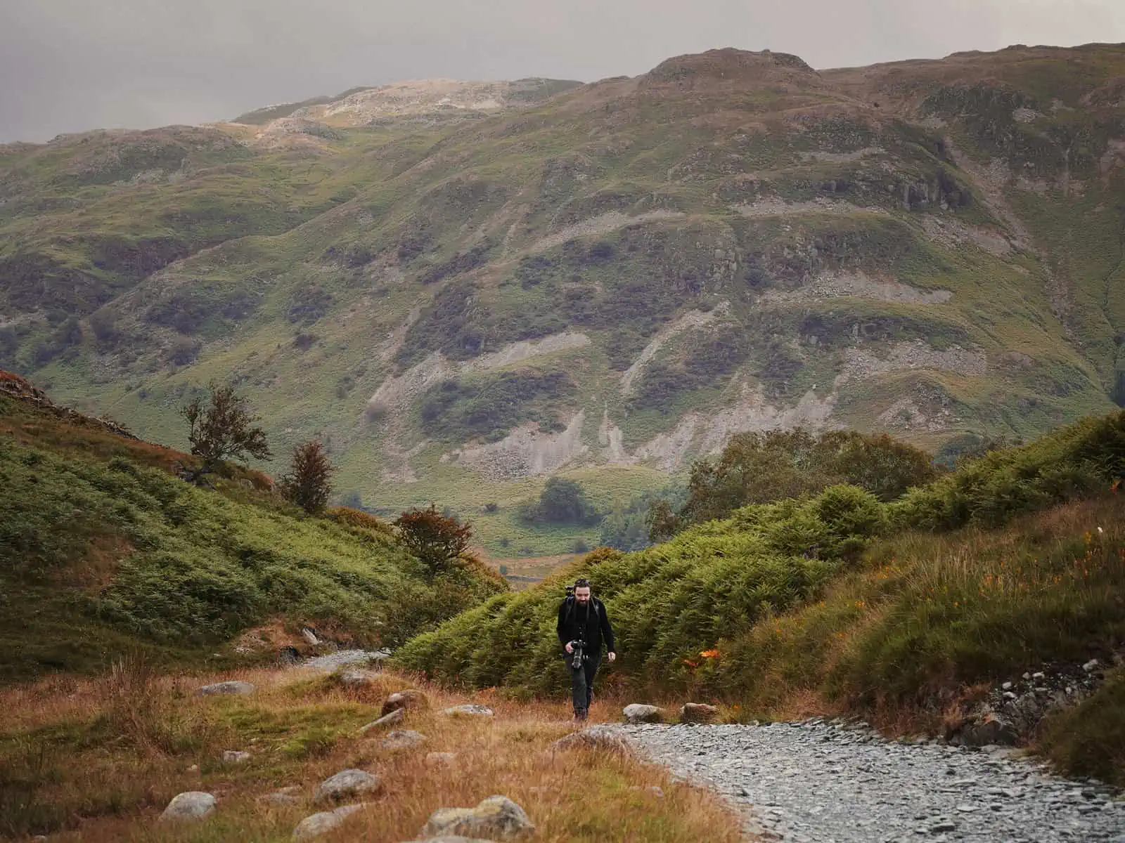 ID: A landscape image. Matt walks up a mountain path and is fairly small in the image as he walks toward the camera. He is mostly wearing black and holds his camera in hand. The mountains in the background are green and brown. The sky is dark grey.