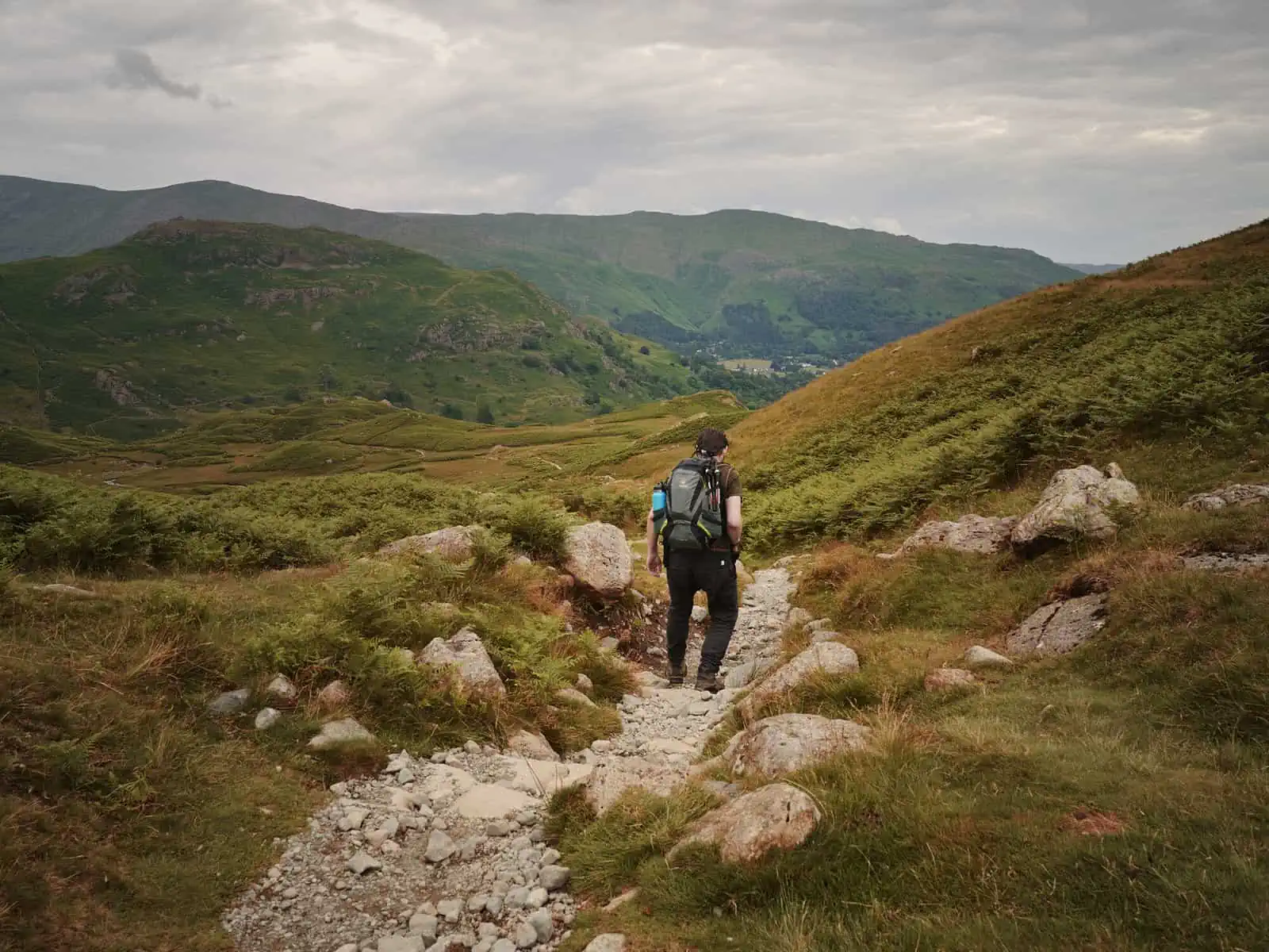 ID: A landscape image. Matt is hiking down a narrow path on a mountain and is centre frame. He is wearing grey trousers, purple top and a grey and green backpack. He is pictured from behind. In the background are more soft mountains with greens and browns from vegetation. The sky is a little overcast.