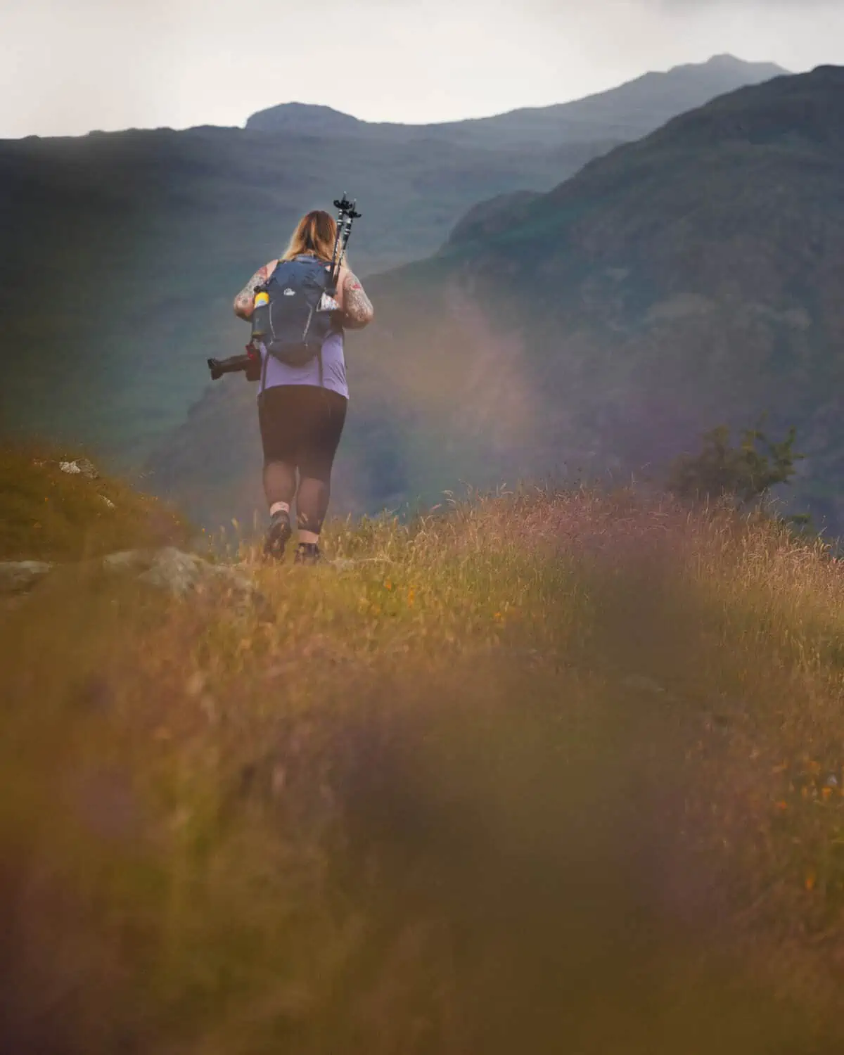 ID: A portrait image. Out of focus foliage takes over the bottom portion of the frame and is made up of orange and yellow hues. In the middle of the frame, Fay is walking away from the camera into a mountain view. Fay is wearing black leggings, purple top, blue backpack and has hiking poles in the side of the pack.