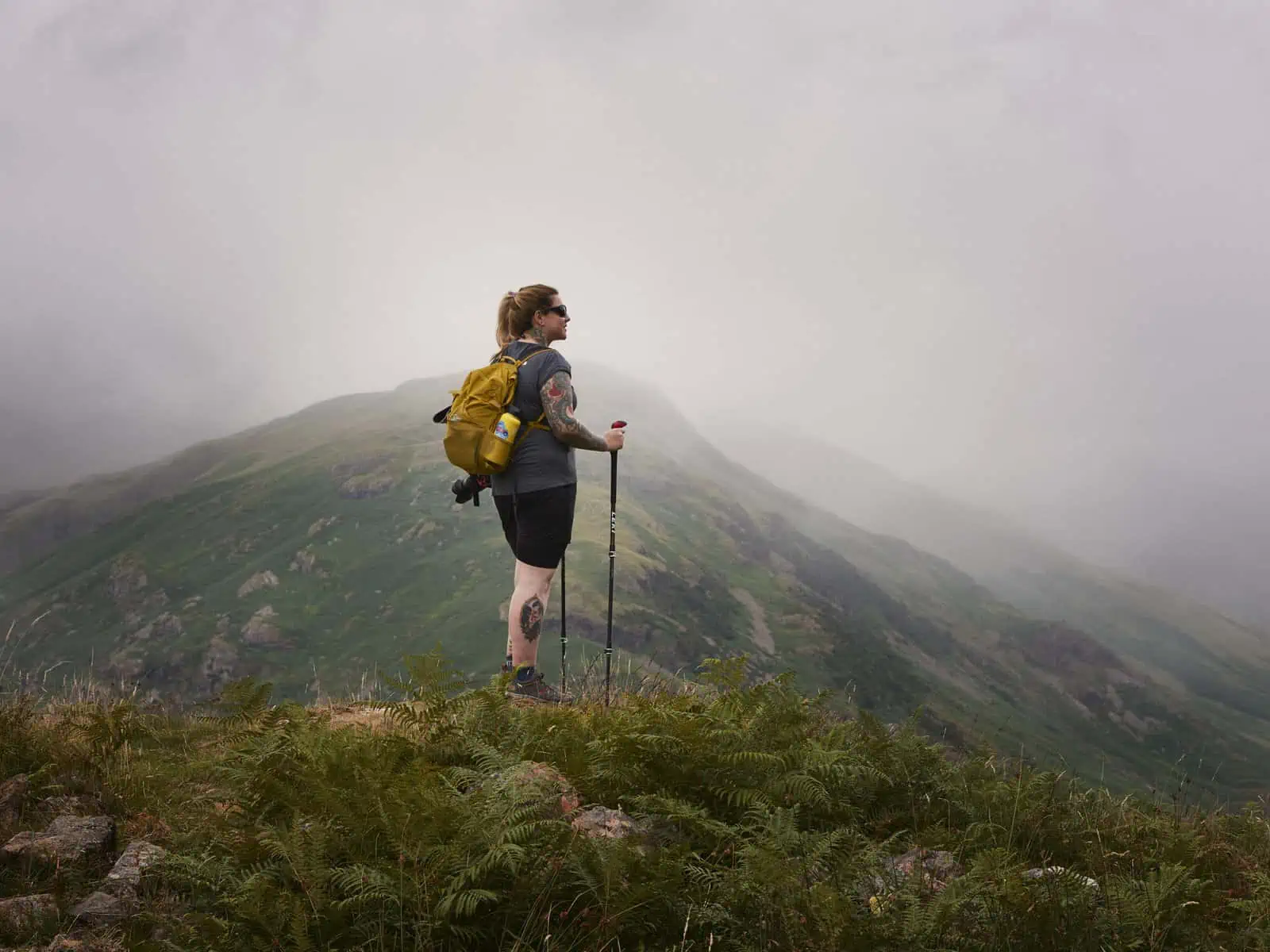 ID: A landscape image. Fay stands on top of a rocky crag looking out at the view. Fay is wearing black shorts, a yellow bag, a grey top and is using hiking poles. The views around are of mountains and it is foggy.&nbsp;