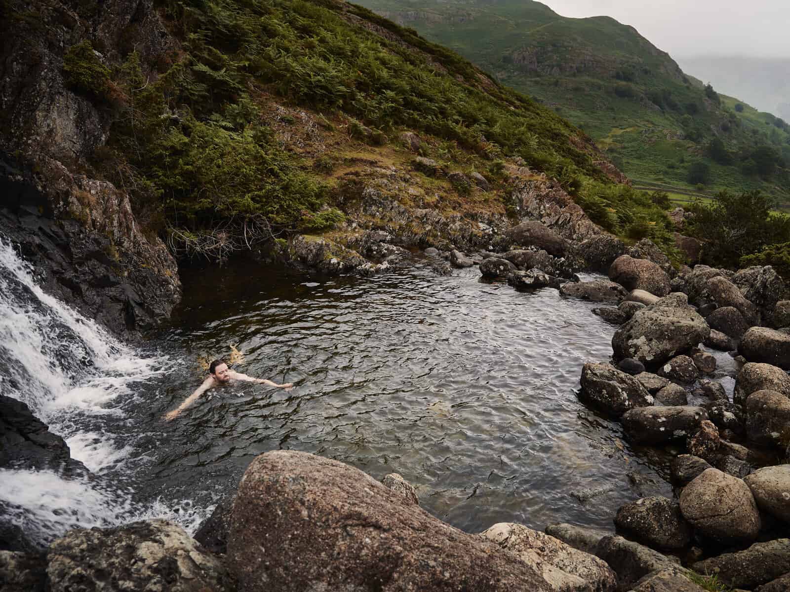 ID: A landscape image. Looking down on Matt who is swimming in a waterfall pool. In the foreground is a waterfall and pool and in the background we can see a mountain view.&nbsp;