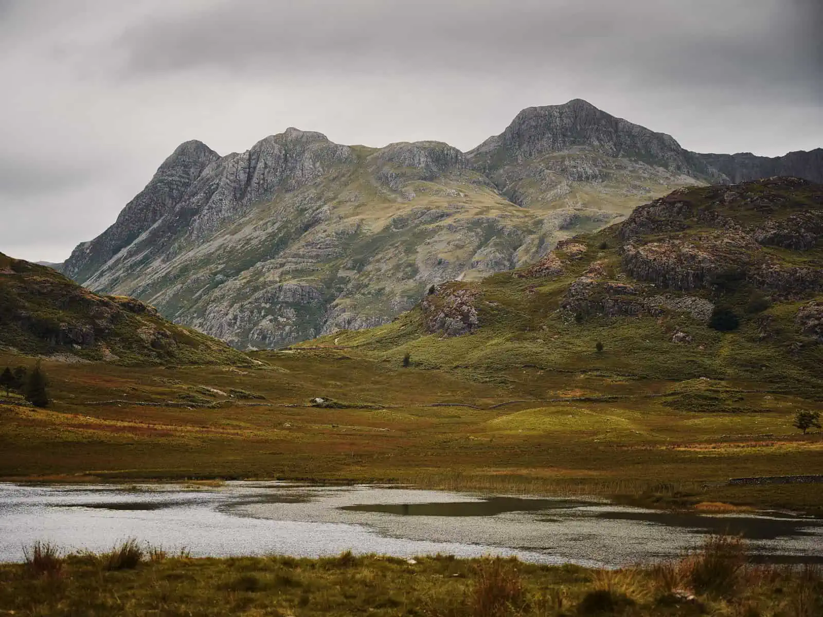 ID: A landscape image. In the foreground is the start of a tarn in the hills – the ground around this is brown and green. As the image progresses, in the background are mountains – rocky and steep. It is overcast.&nbsp;