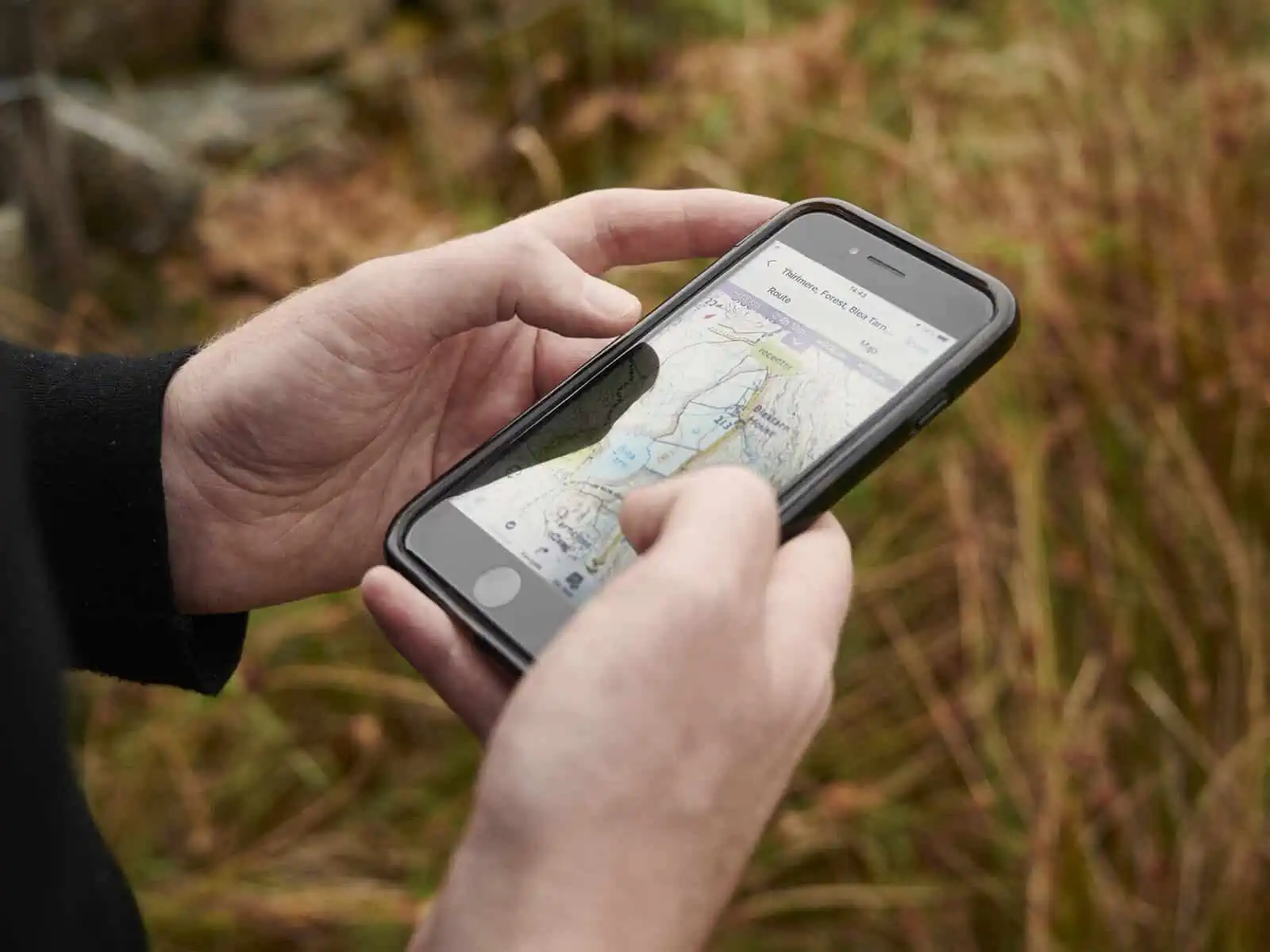 ID: A landscape image. A close up of hands holding a phone looking at a route on Outdooractive. The background is out of focus green and reddish brown grass.&nbsp;