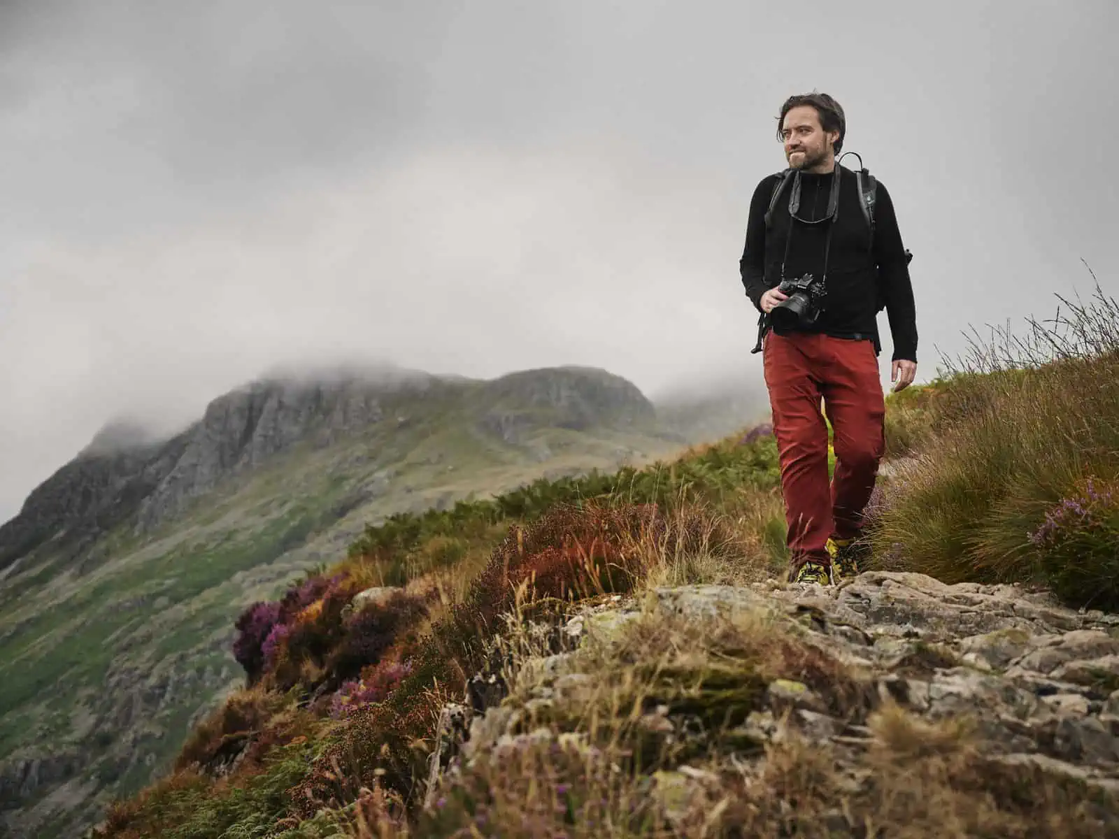 ID: A landscape image. Matt stands to the right of the frame on a rocky high up path. He is wearing a black top and red trousers and has a camera around his neck. There are purple wild flowers around him. In the background are mountains that are bei…