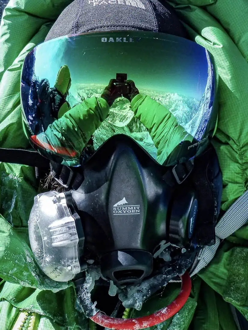 ID: A portrait image. Wearing a high altitude suit with breathing apparatus. In the glasses is a reflection of the arms holding a phone taking a picture with snowy mountains in the background.