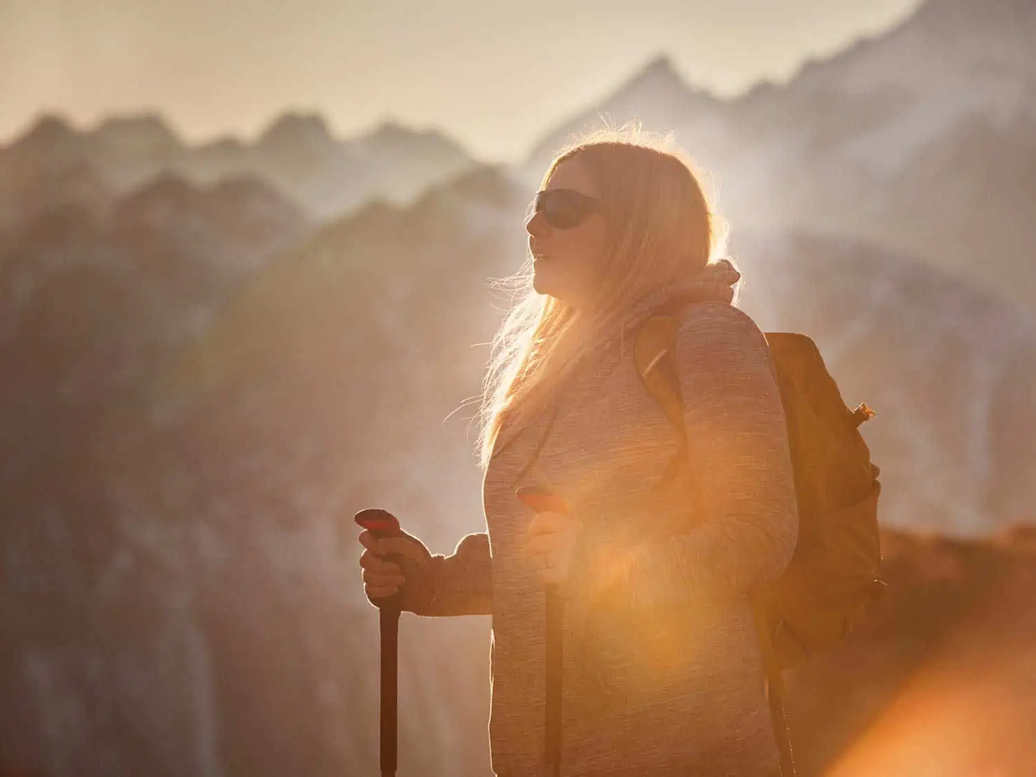 Person wearing grey hoody, yellow bag, black sunglasses with hiking sticks in low level sun with out of focus mountains in background